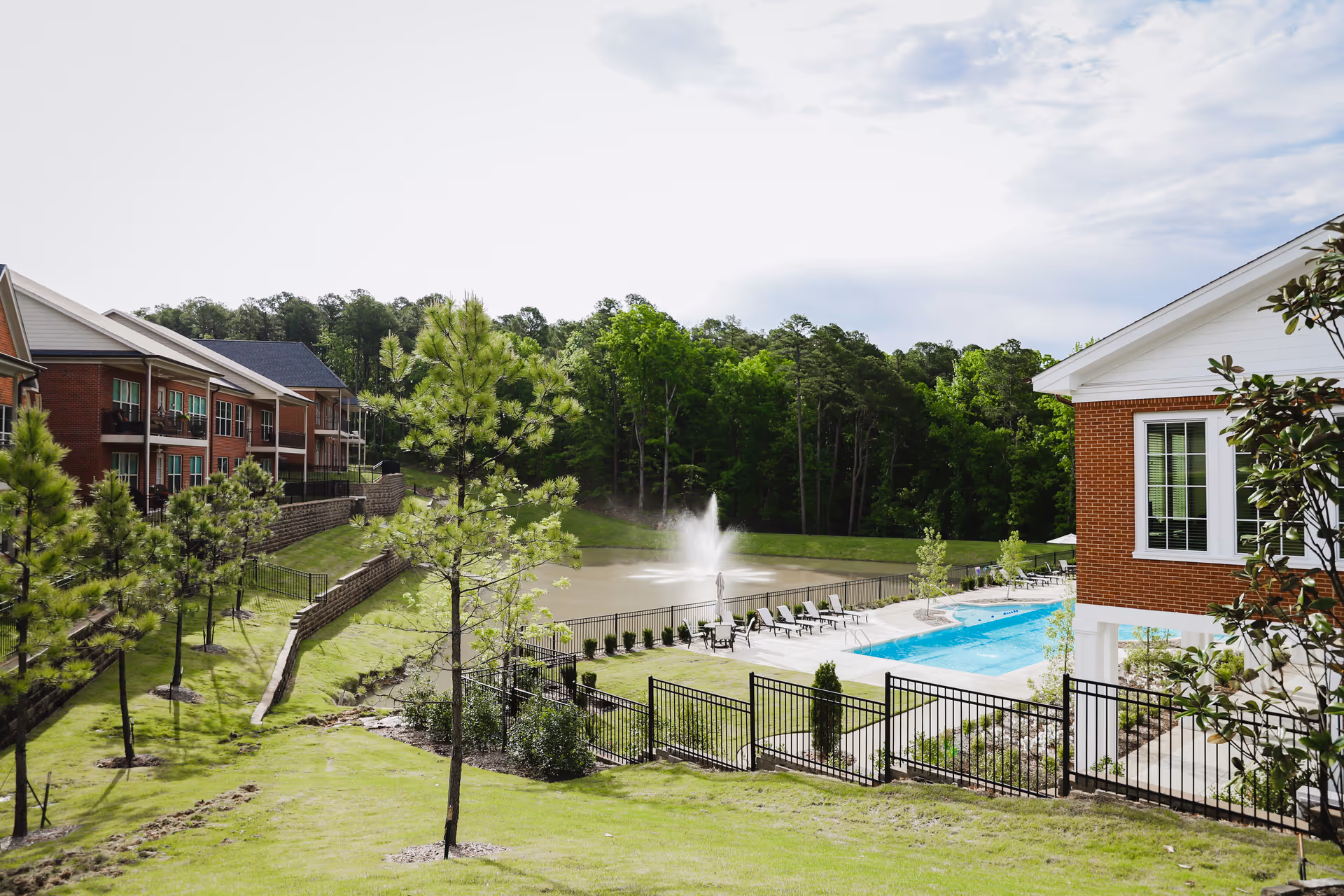 Outdoor view of a senior living campus with brick buildings, a fenced swimming pool, and a fountain in a pond surrounded by trees.