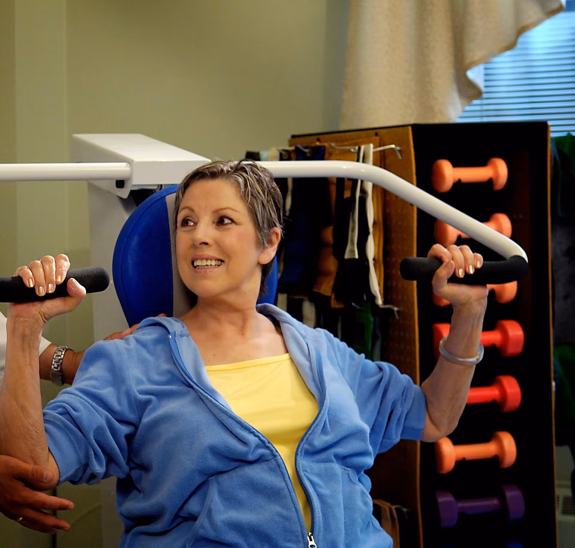 A woman in a blue jacket and yellow top is using a shoulder press exercise machine with assistance from another person. In the background, there is a rack with colorful dumbbells and a window with blinds.