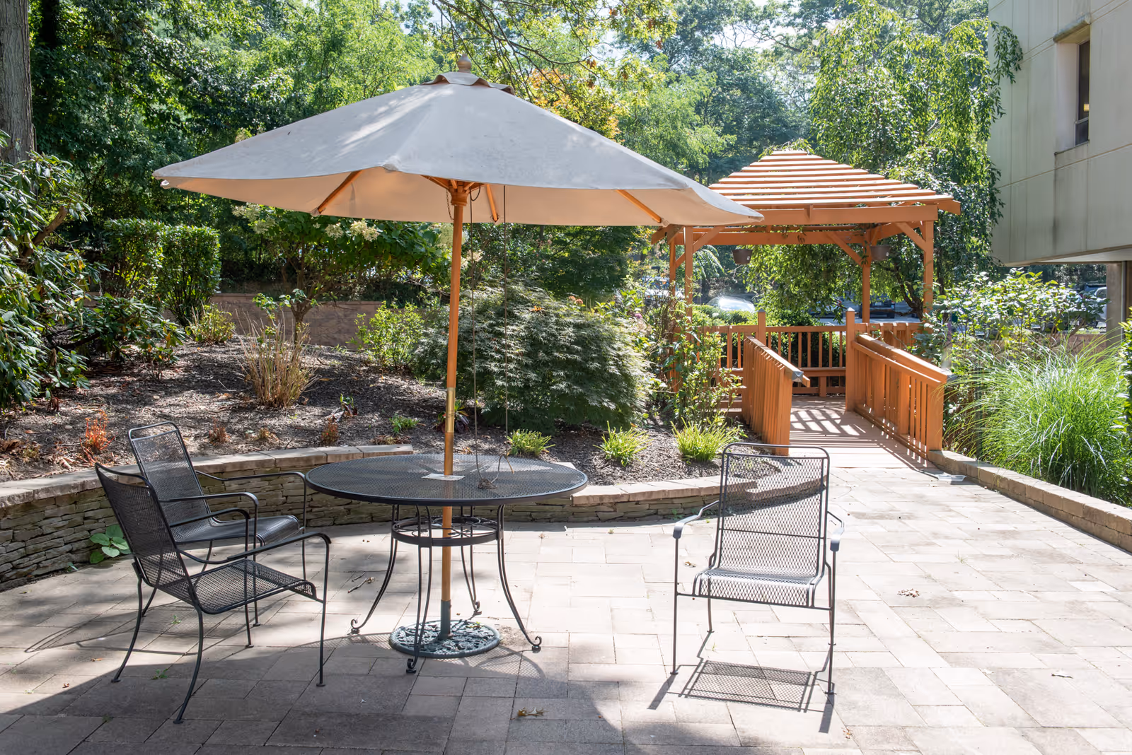 Outdoor patio area with a round metal table and three metal chairs, one of which is positioned under a large beige umbrella. The patio is paved with stone tiles and surrounded by greenery, including bushes and trees. In the background, there is a wooden gazebo with a slatted roof and a small wooden bridge leading to it.