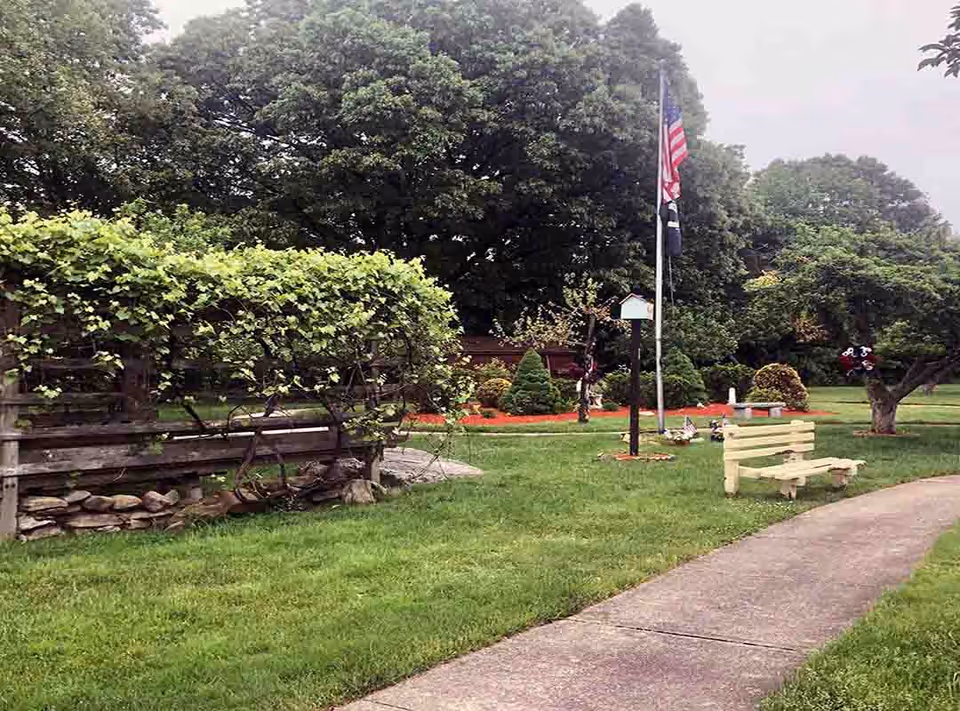 A peaceful outdoor garden area with a paved walkway, green grass, a wooden bench, an American flag on a flagpole, and various trees and shrubs in the background.