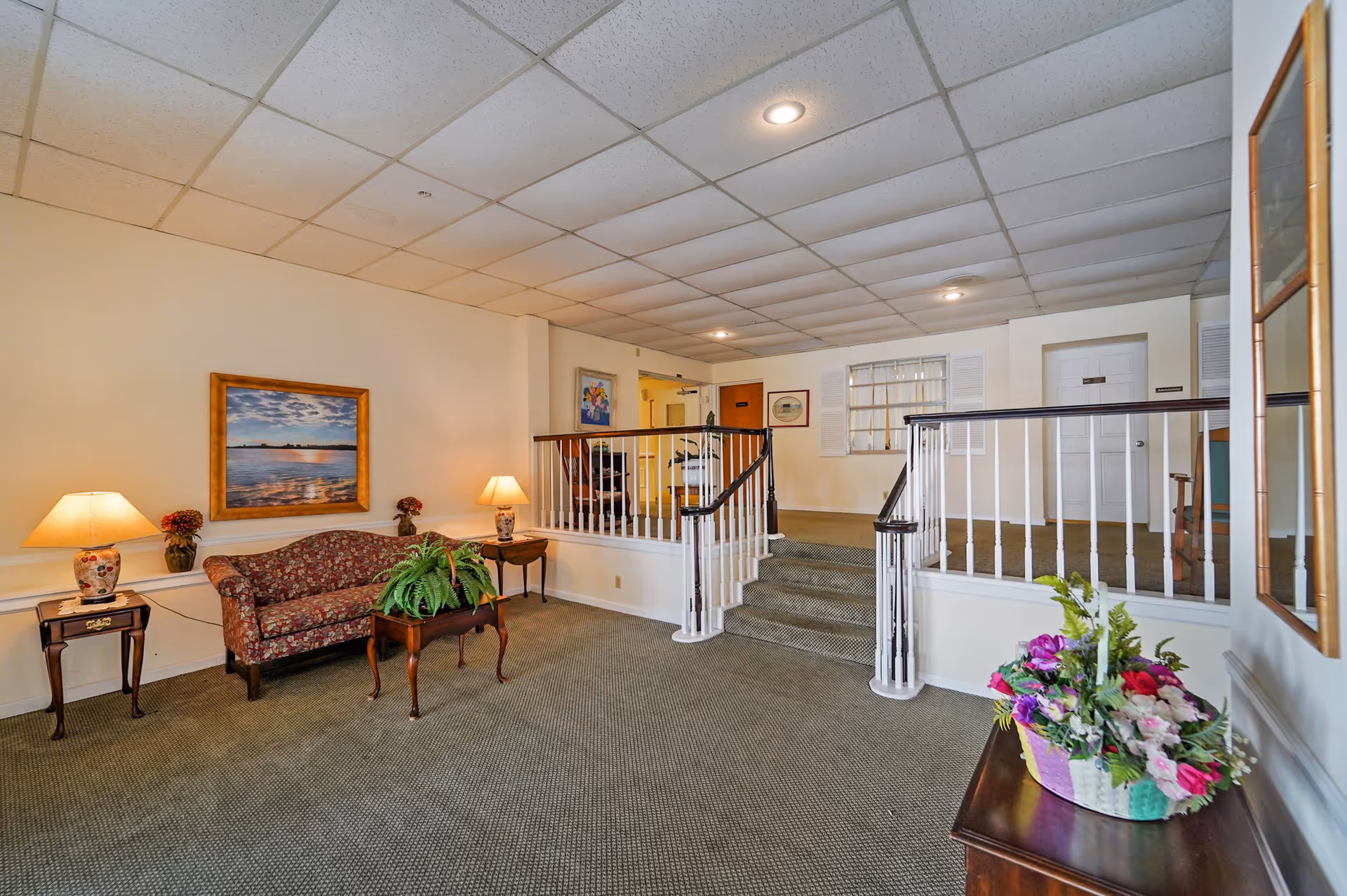 Interior common area with carpeted floor, a floral patterned sofa, two side tables with lamps, a coffee table with a green plant, framed artwork on the walls, and a small staircase with white railings leading to a hallway with doors and additional artwork.