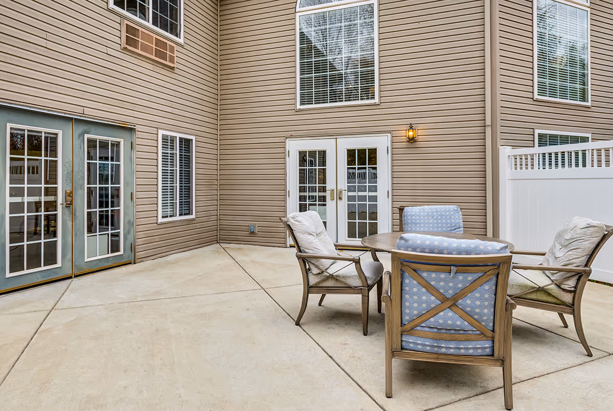 Outdoor patio area with a round table and four cushioned chairs, surrounded by beige siding walls with multiple windows and glass doors. A white privacy fence is visible on the right side.