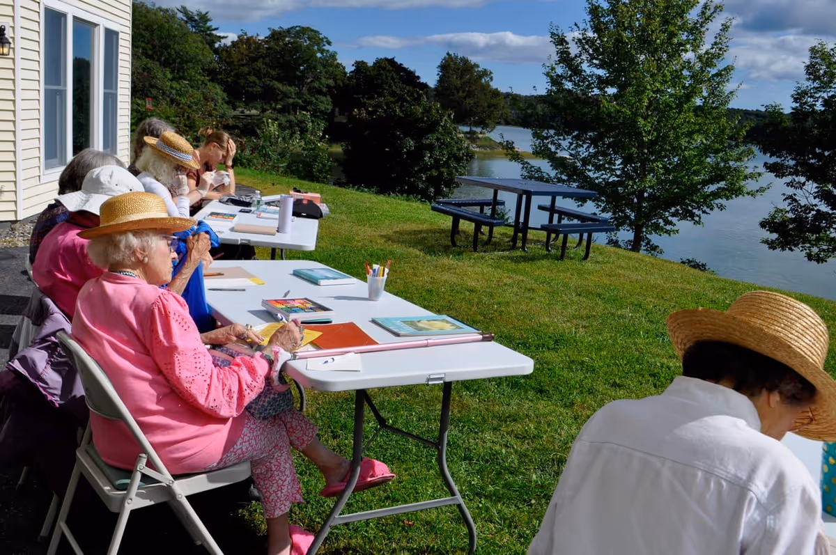 A group of elderly women sitting outdoors at folding tables on a grassy area near a lake, engaged in arts and crafts activities. They are wearing sun hats and light clothing, with art supplies like colored pencils, paper, and books on the tables. Trees and a picnic table are visible in the background under a partly cloudy sky.