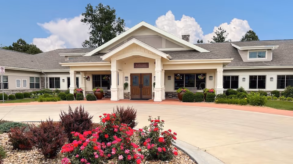 Front exterior view of Lakewood Memory Care facility with a welcoming entrance, double wooden doors, a covered porch supported by columns, and landscaped flower beds with red and pink flowers in the foreground under a partly cloudy sky.