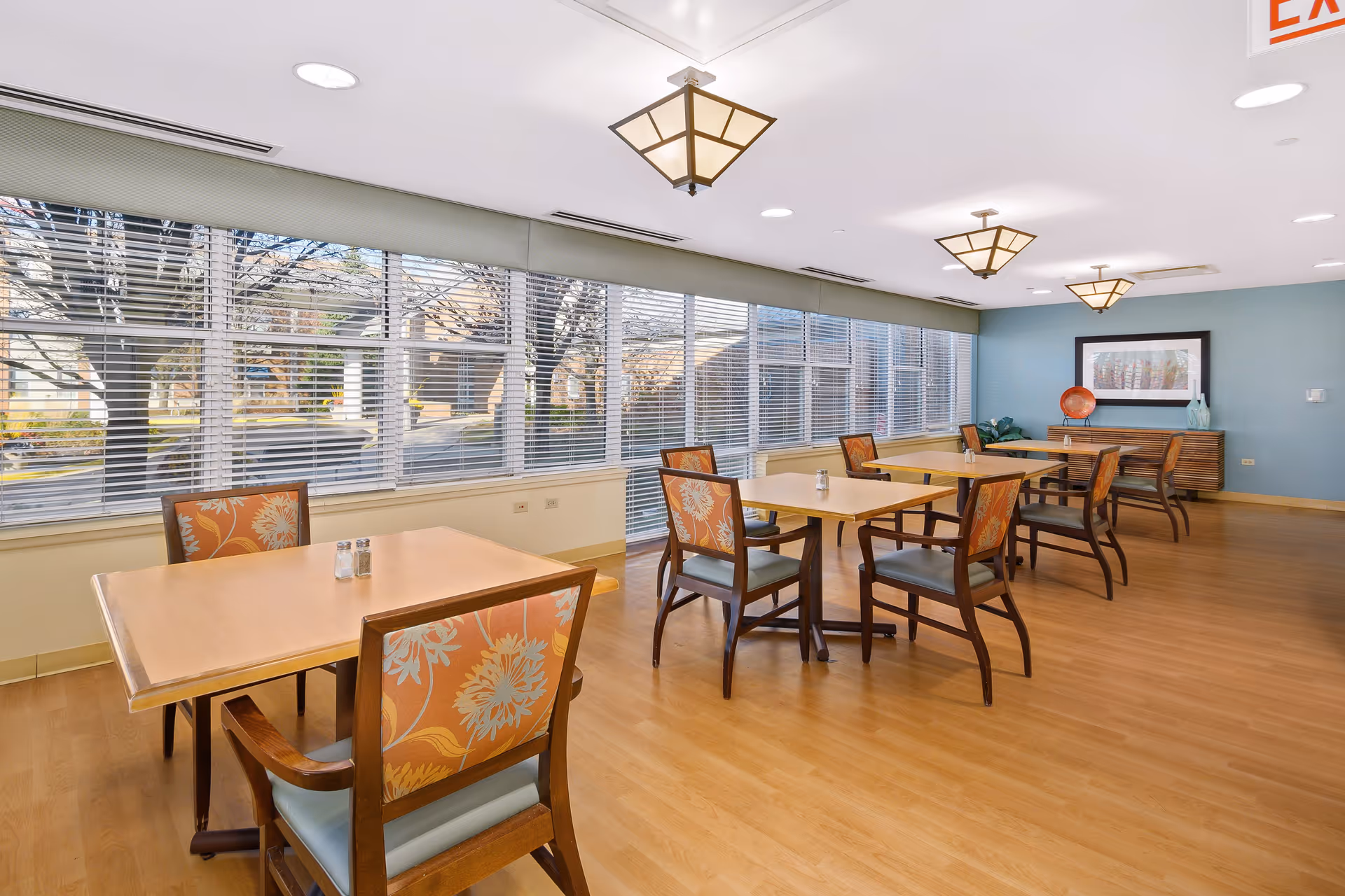 A bright dining room with wooden floors and several tables, each surrounded by chairs with floral patterned upholstery. Large windows with blinds line one wall, letting in natural light. The room has modern ceiling lights and a blue accent wall with framed artwork and decorative items on a sideboard.