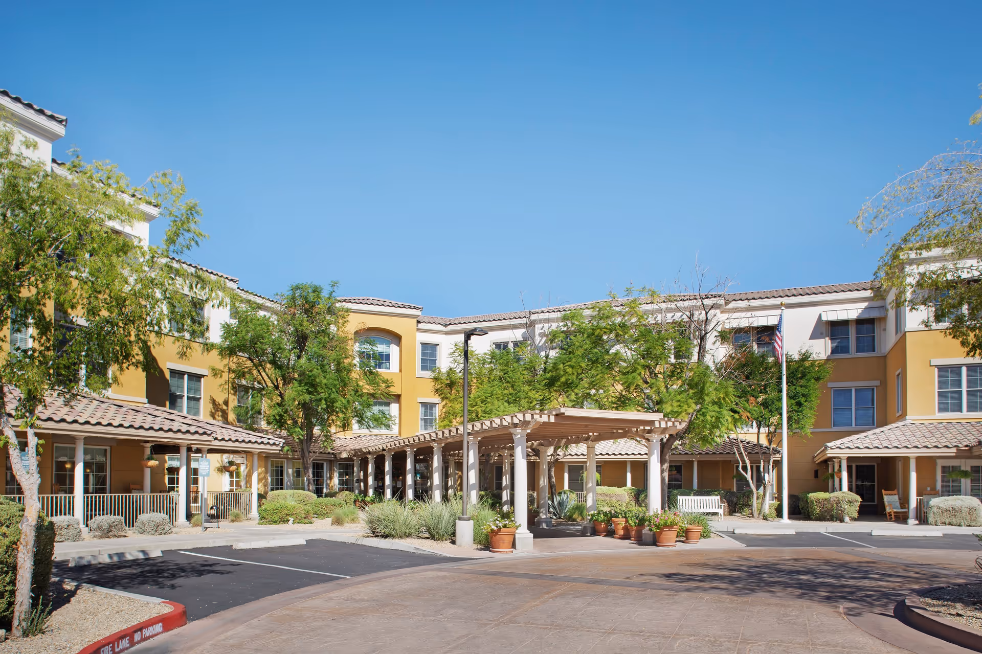 Exterior view of Sunrise of Scottsdale senior living facility showing a three-story building with yellow and white walls, tiled roofs, trees, shrubs, and a covered entrance area with potted plants under a clear blue sky.