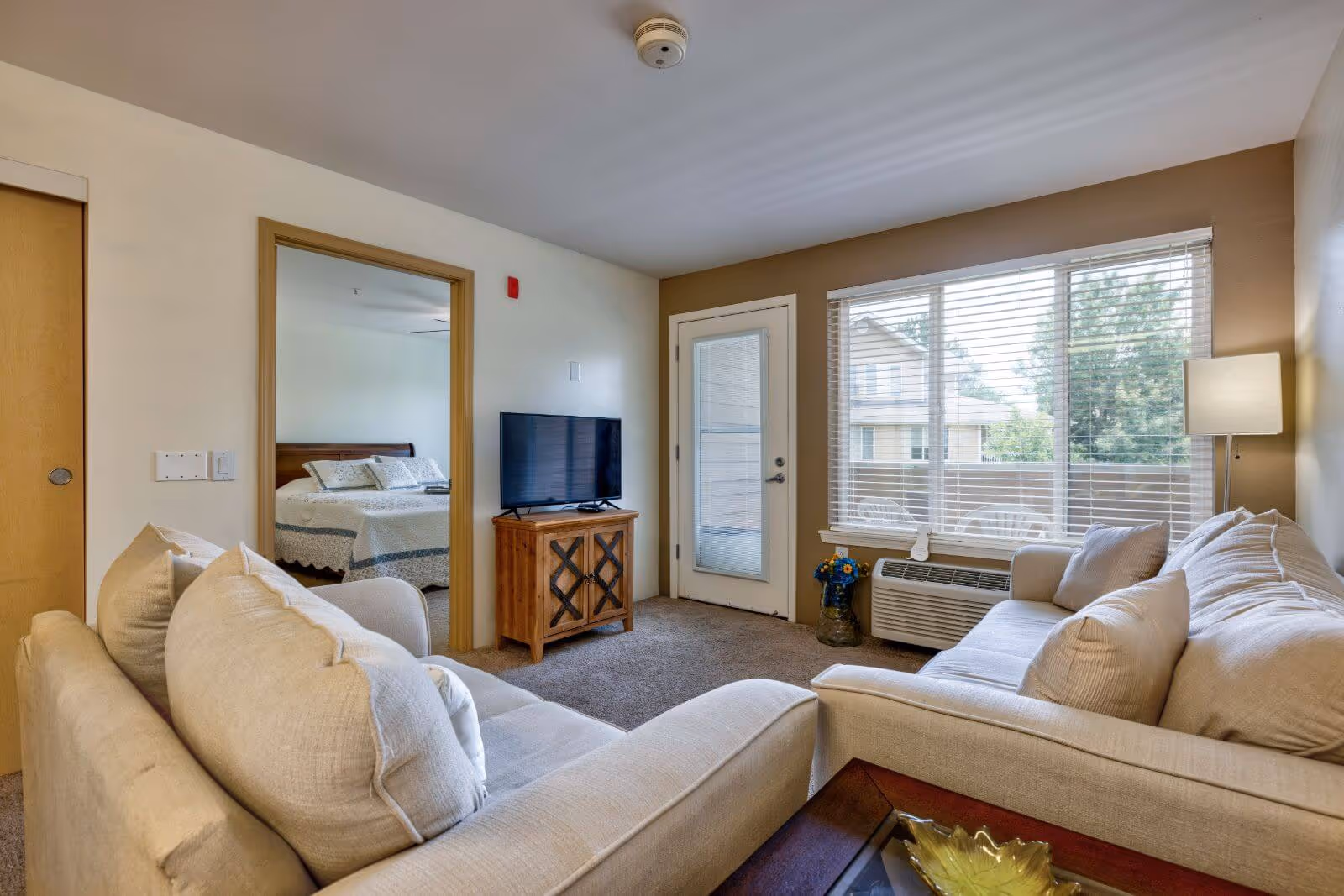Bright living room with beige sofas facing a TV, a large window and a door to a balcony, with a view into an adjacent bedroom.