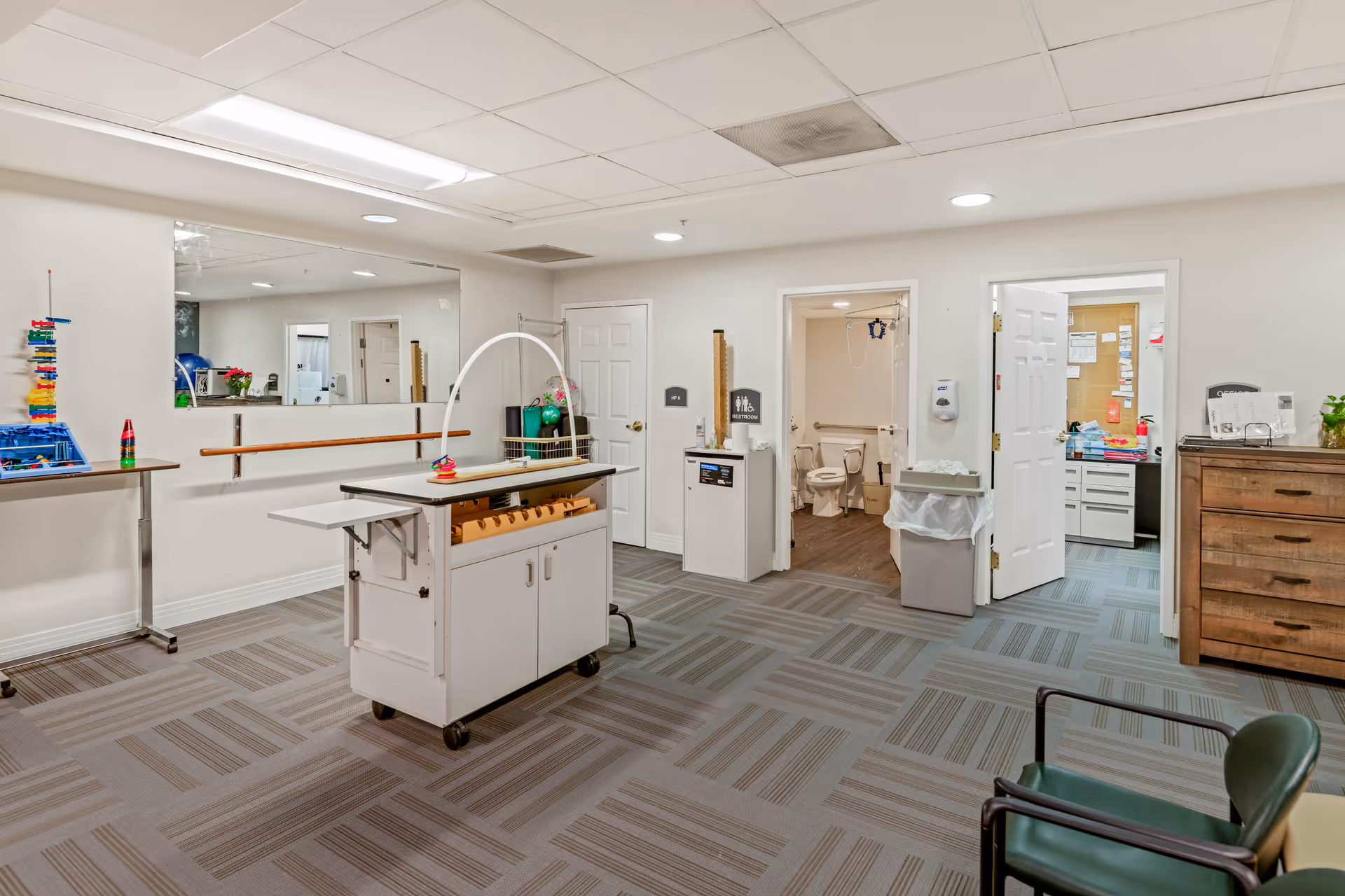 Bright multipurpose therapy/activity room with a rolling cart, mirrors, handrails, seating, and open doors to adjacent rooms.