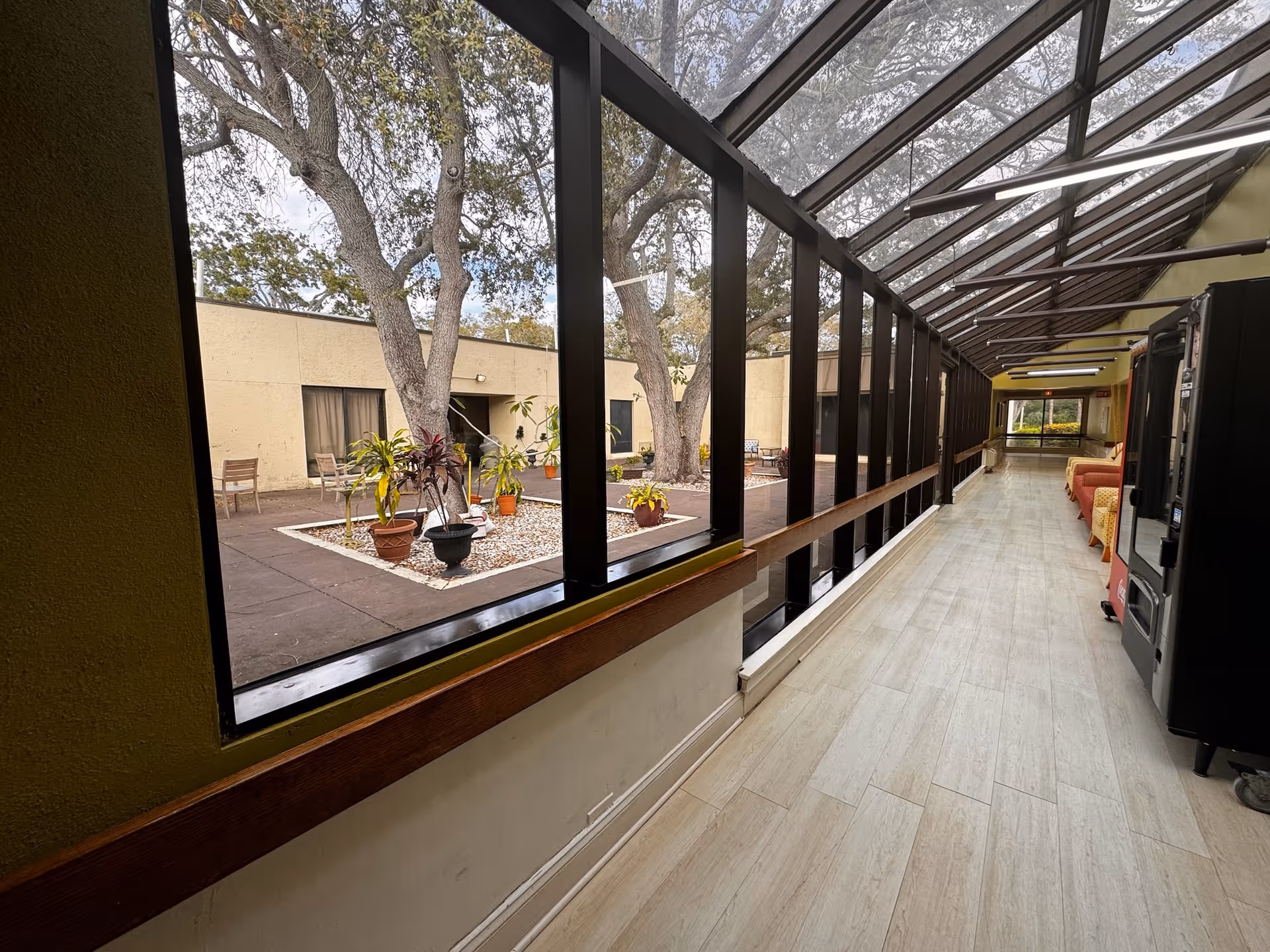 A long indoor hallway with large windows on the left side overlooking an outdoor courtyard with trees and potted plants. The hallway has light-colored flooring, a vending machine on the right, and seating areas with chairs and couches further down the corridor. The ceiling is slanted with glass panels allowing natural light to enter.