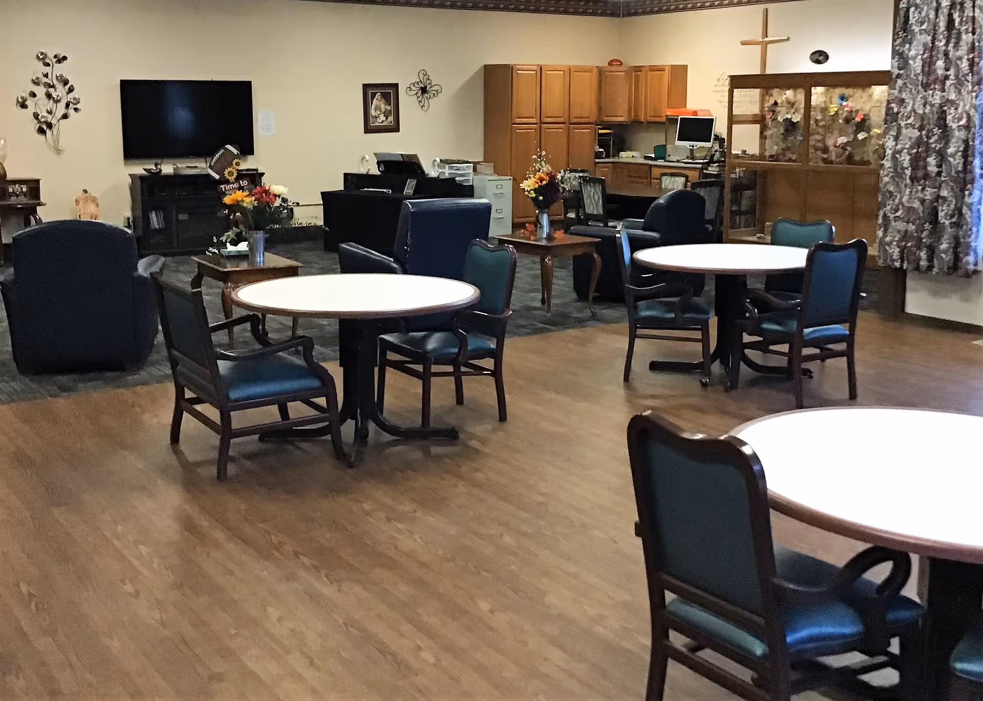 Communal activity/dining room with round tables and chairs, lounge seating, a TV, and a reception/office area.