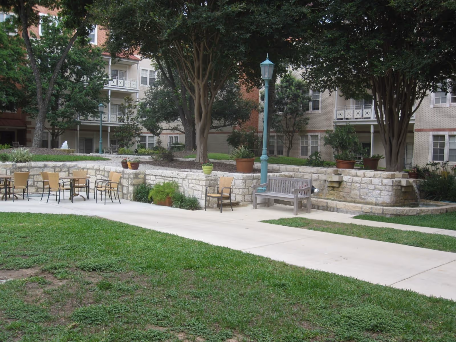 Outdoor courtyard area with green grass, a paved walkway, several chairs and tables, a wooden bench, potted plants, trees, a stone wall with water features, and a multi-story building with balconies in the background.