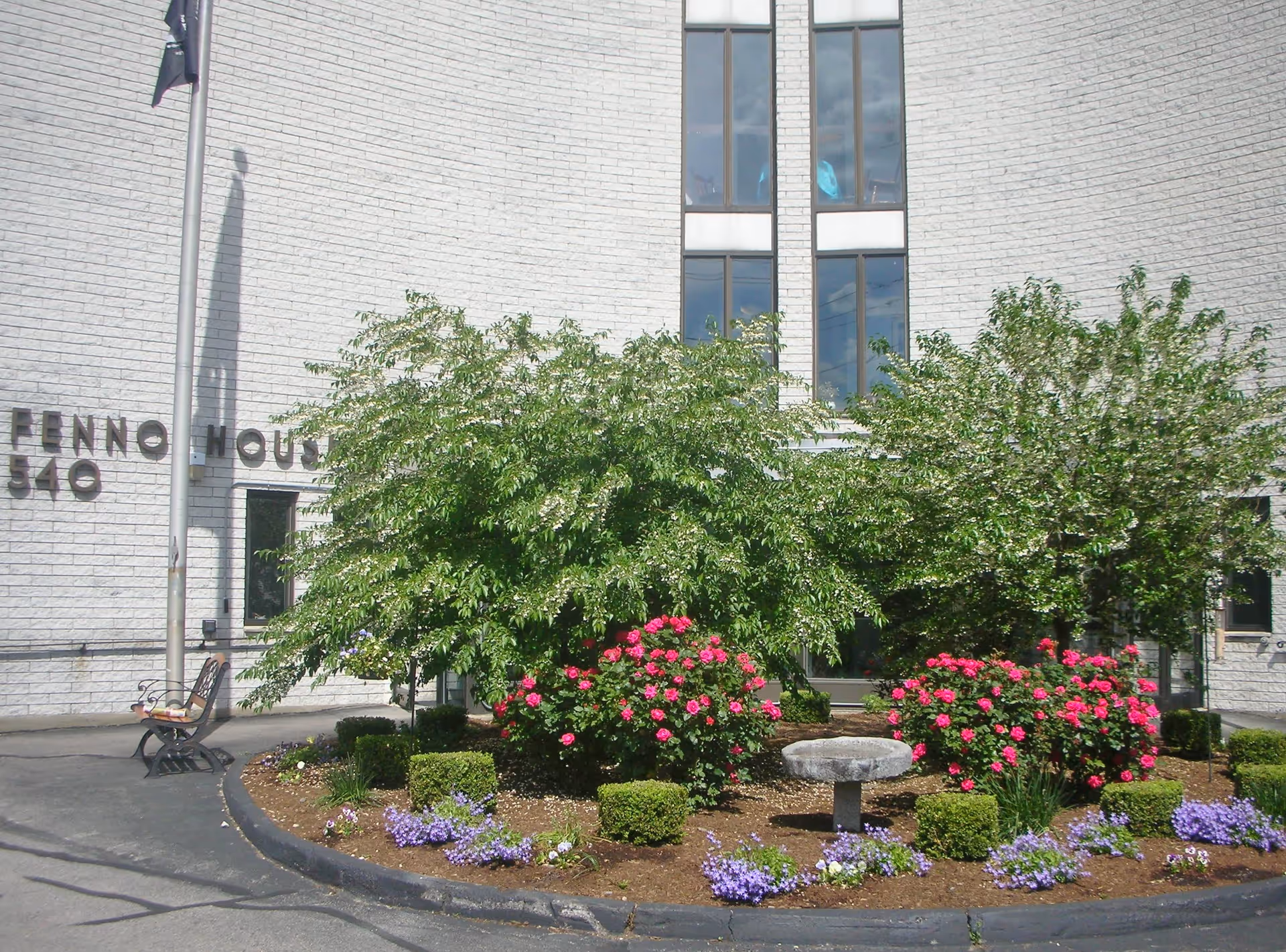 Front exterior view of Fenno House building with a curved white brick wall, tall windows, a flagpole, a bench, and a landscaped garden featuring green bushes, pink flowers, and purple flowers.