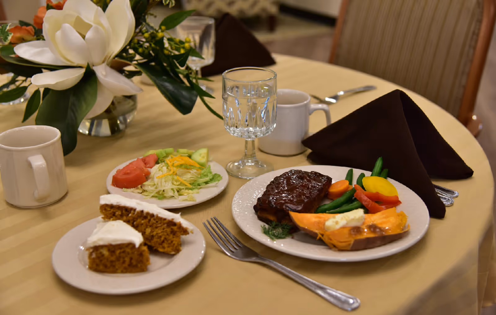 A dining table set with a meal including a plate of ribs with mixed vegetables and a baked sweet potato, a side salad with lettuce, tomato, and cucumber, two slices of carrot cake with white frosting on a separate plate, a glass of water, two white coffee mugs, and a dark brown folded napkin. A floral centerpiece with white flowers and green leaves is also on the table.