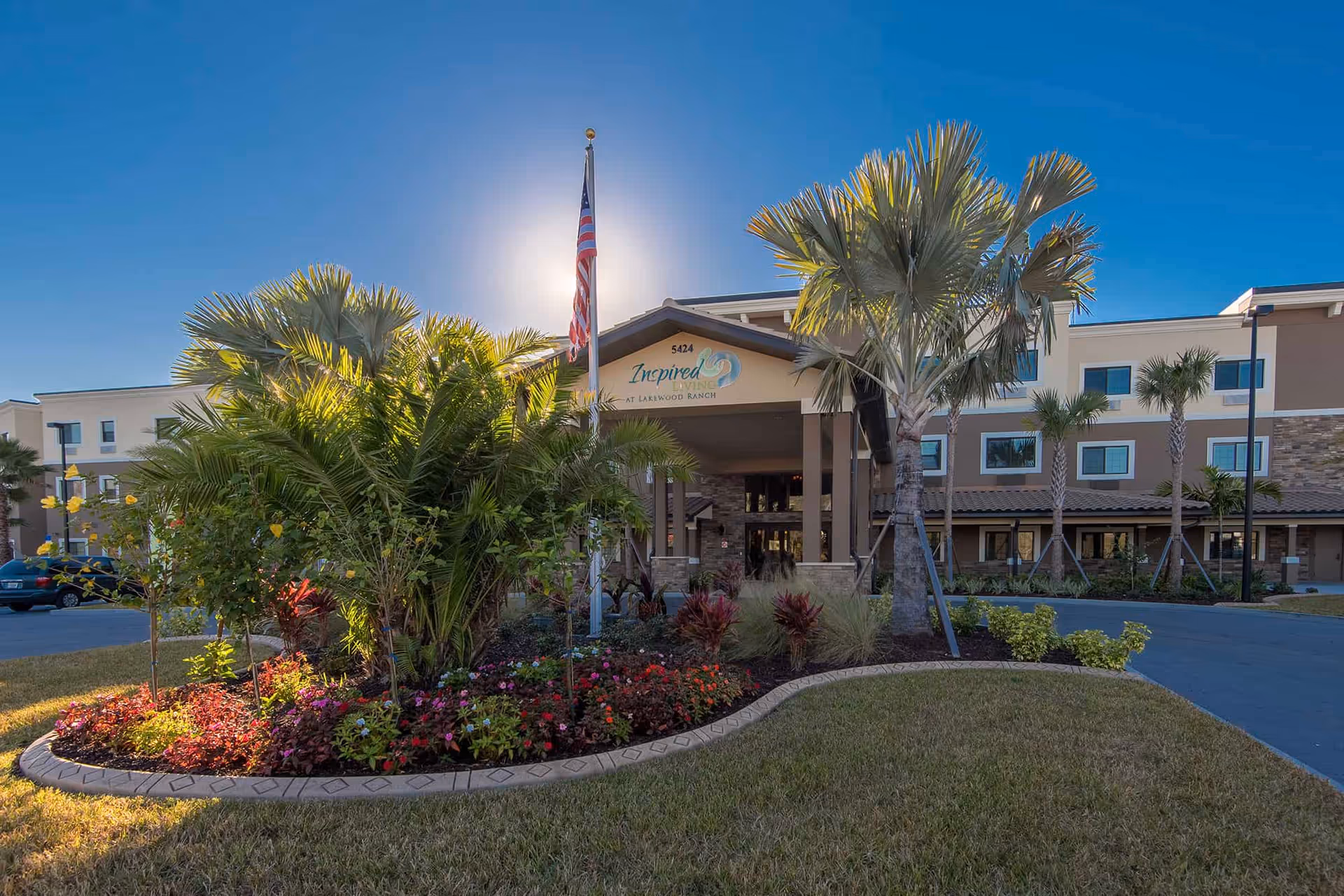 Exterior view of Inspired Living Lakewood Ranch senior living facility with a landscaped garden, palm trees, an American flag, and a clear blue sky.