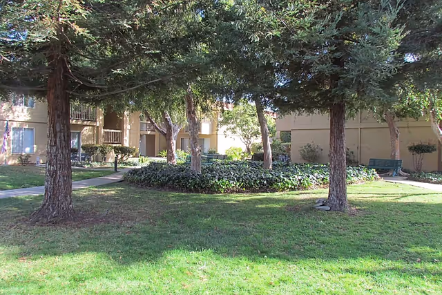 Shaded grassy courtyard with trees, a circular landscaped planter, benches and the exterior entrances of a retirement community building.