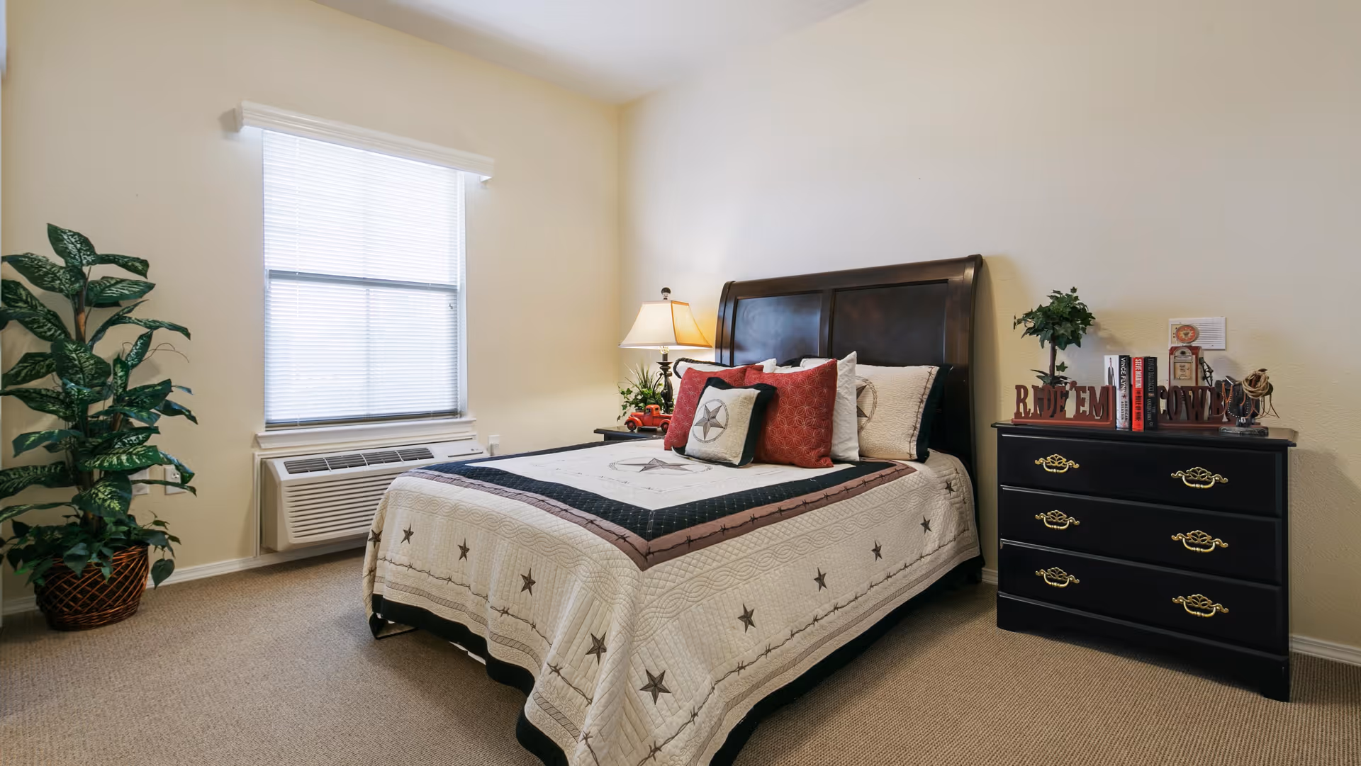 A neatly made bed with a dark wooden headboard in a bedroom. The bed has a quilted bedspread with star patterns and several decorative pillows. To the right of the bed is a black dresser with gold handles, decorated with books, a small plant, and decorative items. A table lamp with a beige shade is on a nightstand next to the bed. A large window with closed blinds is on the left wall, and a potted plant is placed on the floor near the window. The walls are light-colored, and the floor is carpeted.