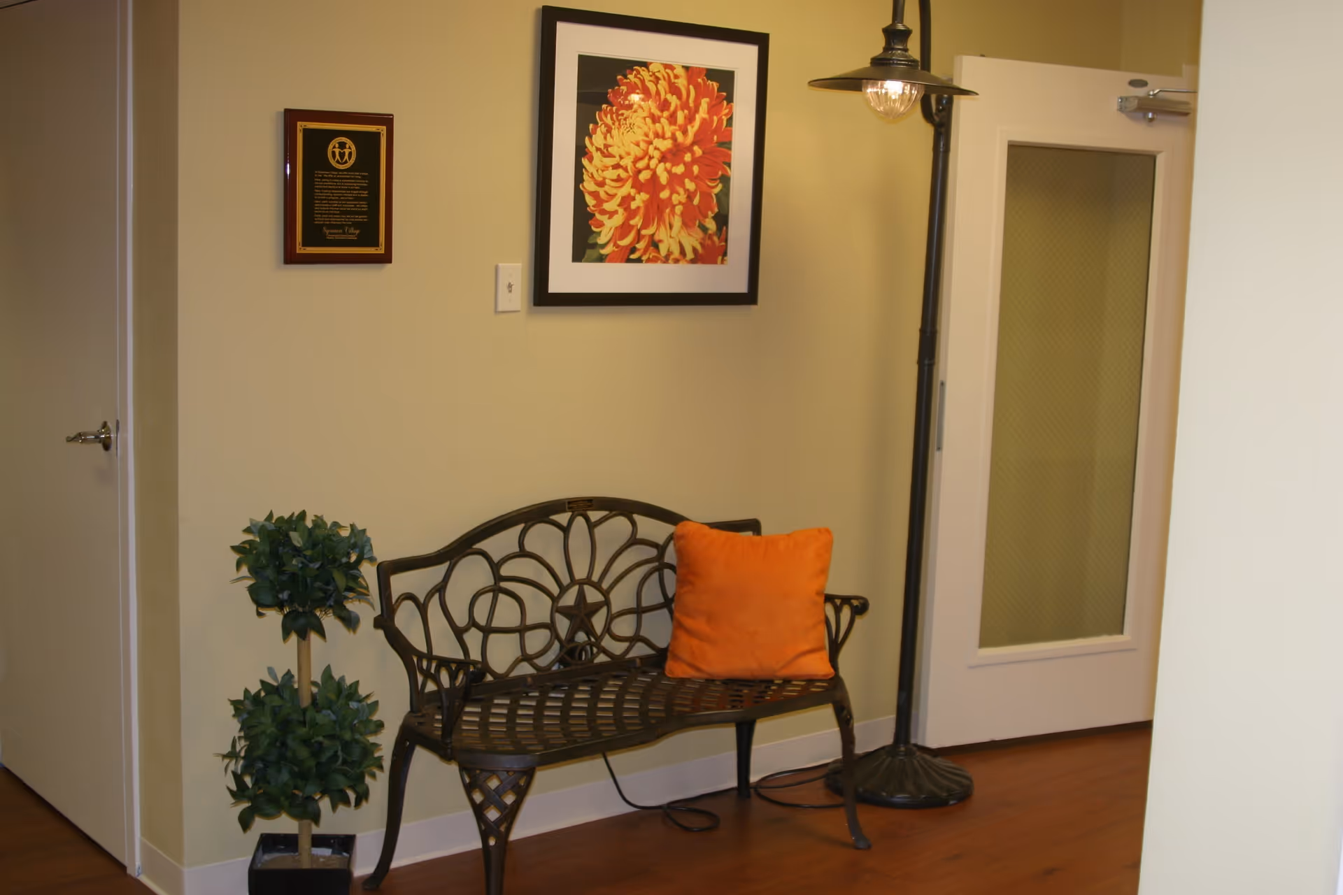 An interior corner of a room with a decorative metal bench featuring an orange cushion, a tall floor lamp with a visible light bulb, a framed picture of a large orange and yellow flower on the wall, a small potted plant with two green leafy sections, and a closed white door with a window panel.