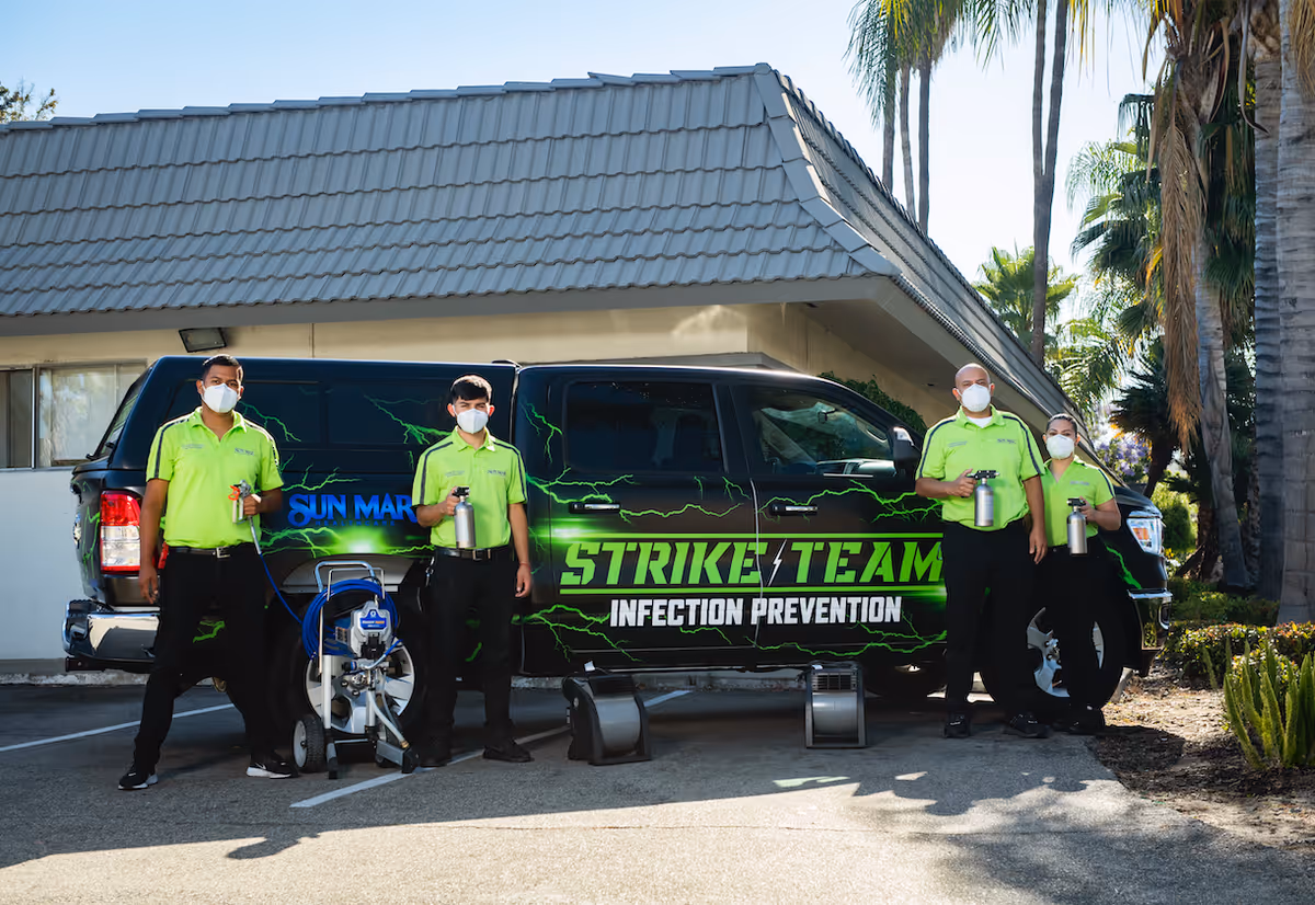 Four people wearing bright green shirts and black pants stand in front of a black vehicle with green lightning graphics and the text 'STRIKE TEAM INFECTION PREVENTION'. They are all wearing white face masks and holding spray bottles. The setting appears to be an outdoor parking area next to a building with a tiled roof and palm trees in the background.