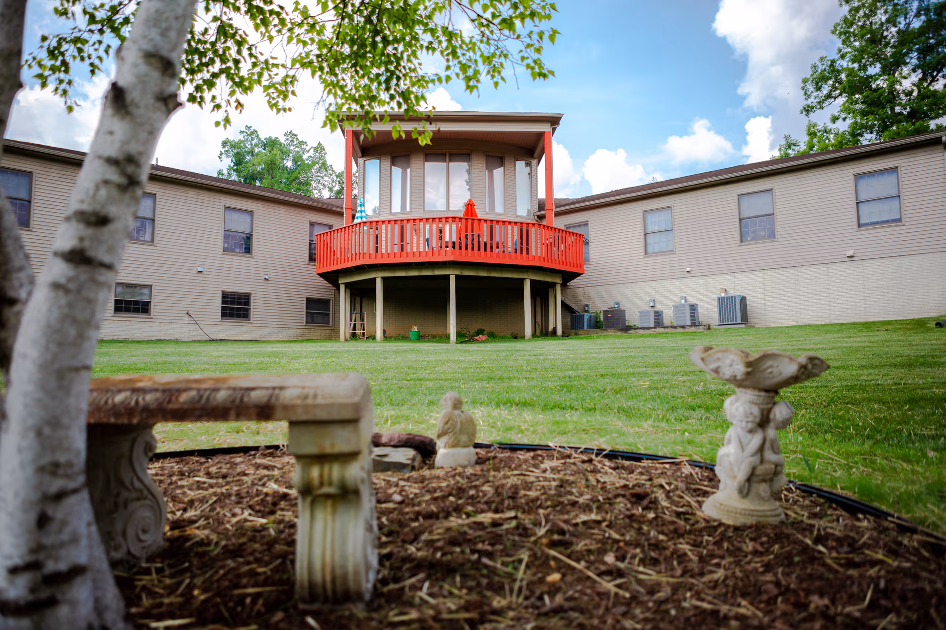 View of the exterior of a beige two-story building with a red balcony in the center. In the foreground, there is a small garden area with a stone bench, a tree, and decorative statues on a mulched bed. The sky is partly cloudy with blue patches visible.