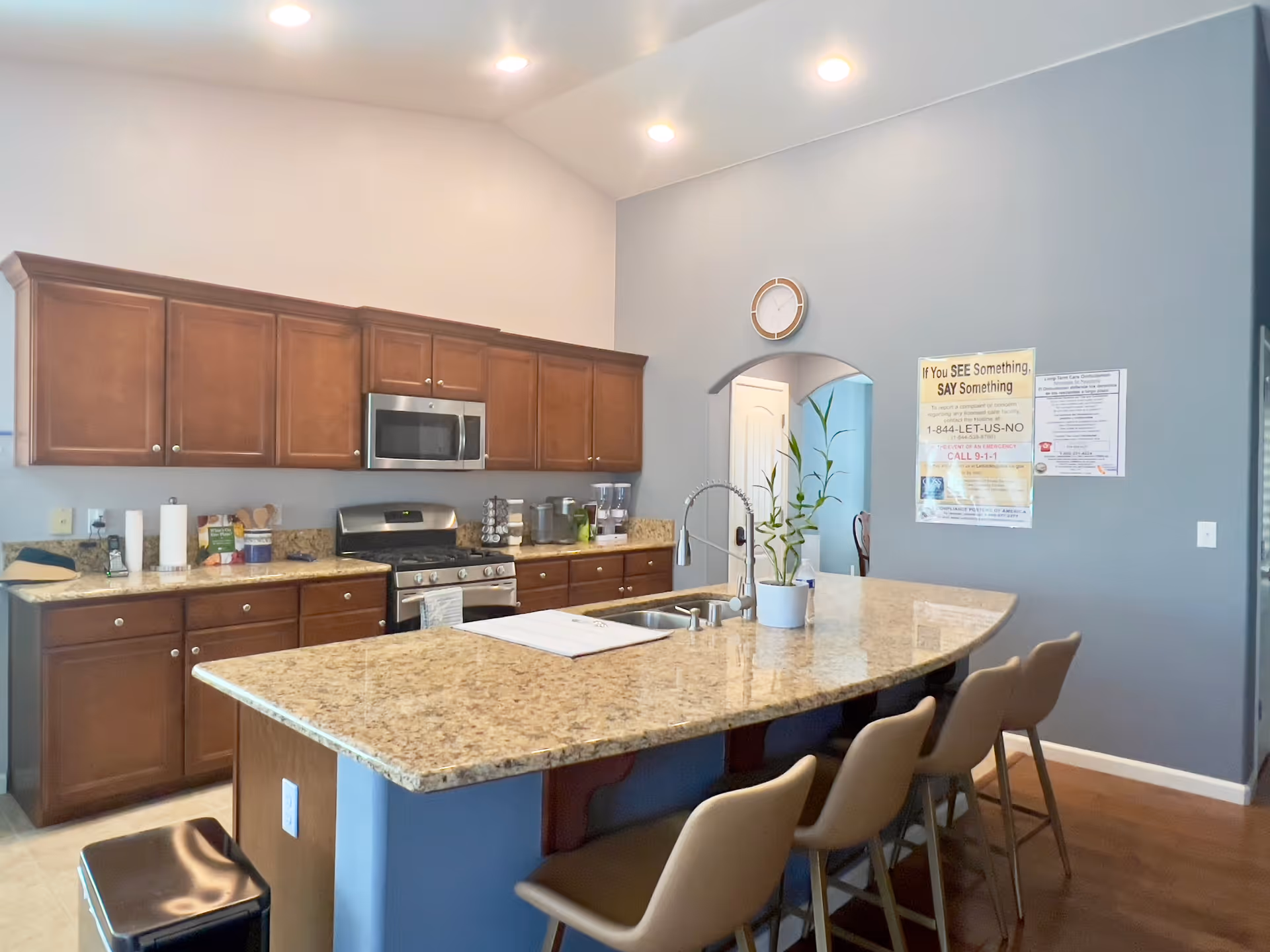 A modern kitchen with wooden cabinets, a granite countertop island with a built-in sink, four beige bar stools, a stainless steel microwave and stove, and a small potted plant on the island. The walls are painted light gray, and there are informational posters on the wall near an arched doorway.