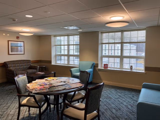 A cozy senior living common area with a round table covered in magazines surrounded by four chairs. There are two large windows letting in natural light, a patterned couch, a blue armchair, and a blue sofa. The walls are beige with a framed picture hanging, and the ceiling has recessed and circular lights.