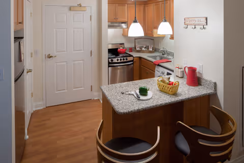 A small kitchen area with wooden cabinets, granite countertops, and stainless steel appliances including a stove and microwave. Two pendant lights hang above a breakfast bar with two wooden chairs. On the counter, there is a basket with fruit, a red pitcher, and a plate with a green bell pepper and utensils. A white door is visible in the background, and a wall sign reads 'Worry is...'.
