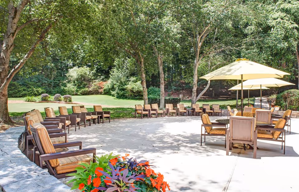 Outdoor patio area with multiple cushioned chairs arranged in a semicircle around a paved space, surrounded by trees and greenery. There are also tables with yellow umbrellas providing shade. Colorful flowers are visible in the foreground.