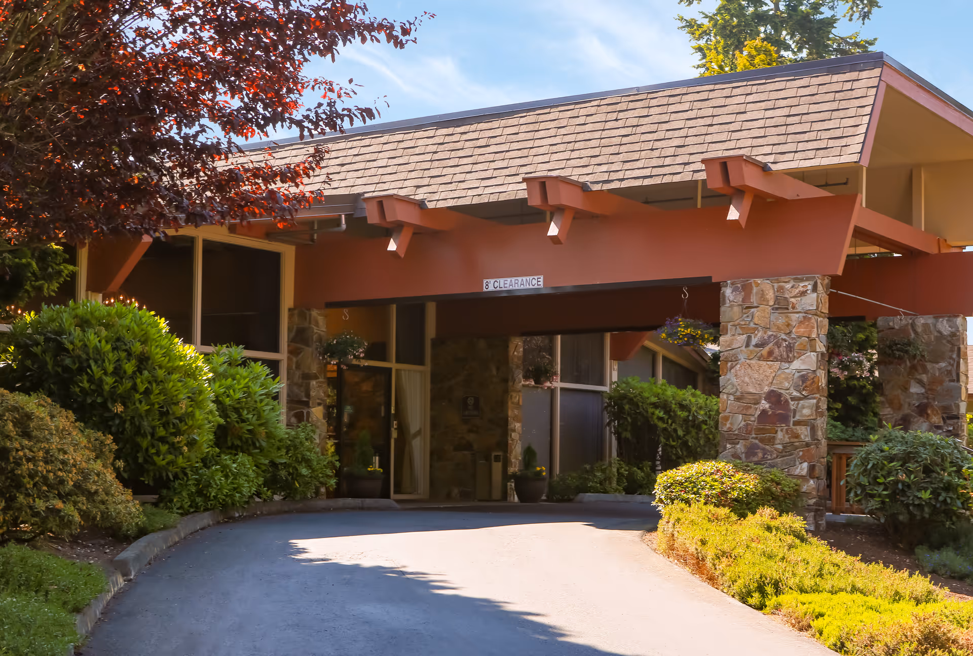 Covered driveway entrance with stone pillars and landscaped shrubs in front of a senior care building.