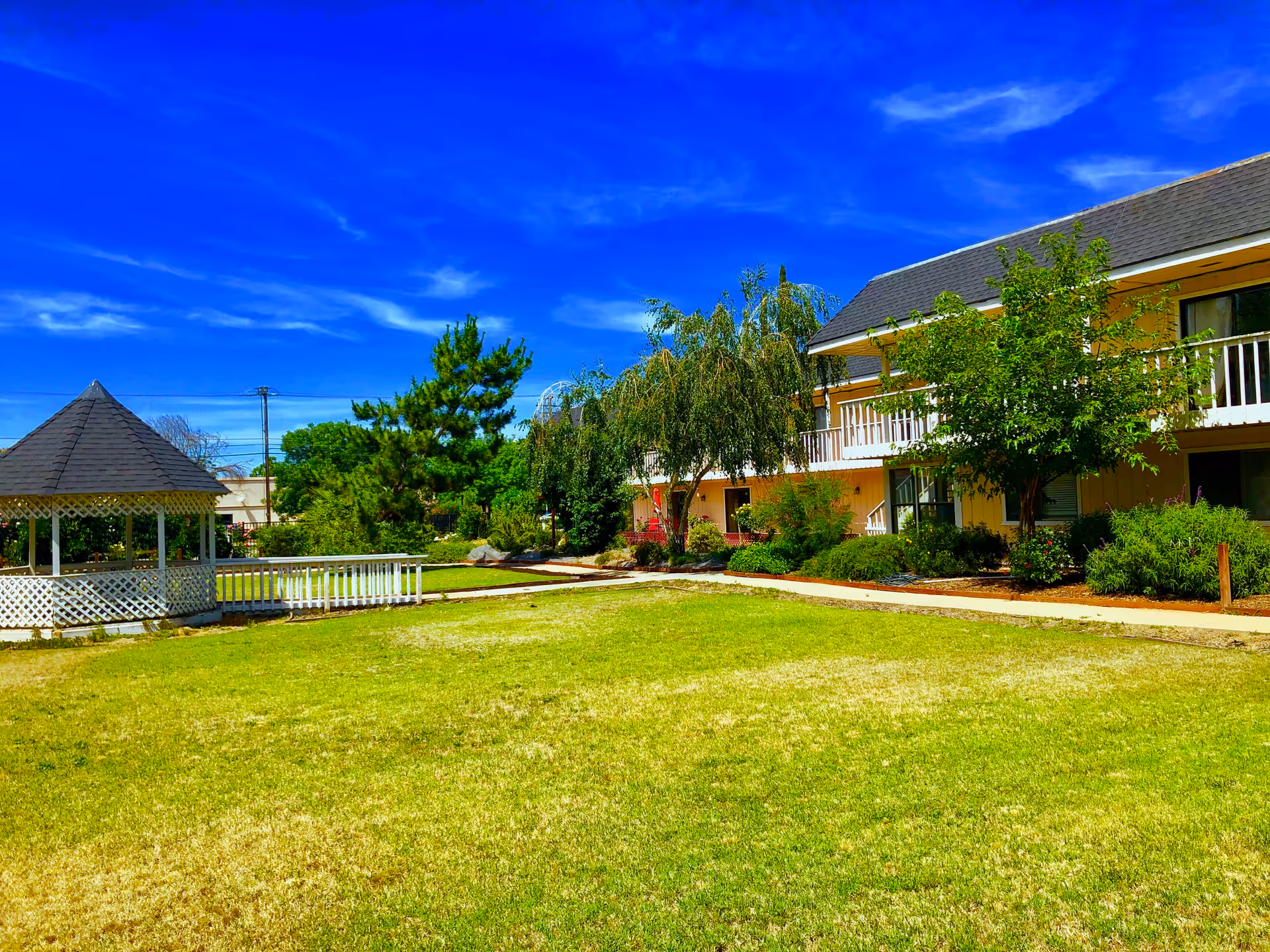 A bright outdoor scene at St. Francis Assisted Care featuring a well-maintained grassy lawn, a white gazebo with a dark shingled roof on the left, and a two-story yellow building with white railings and balconies on the right. Trees and shrubs are planted along the building and pathways, under a clear blue sky with wispy clouds.