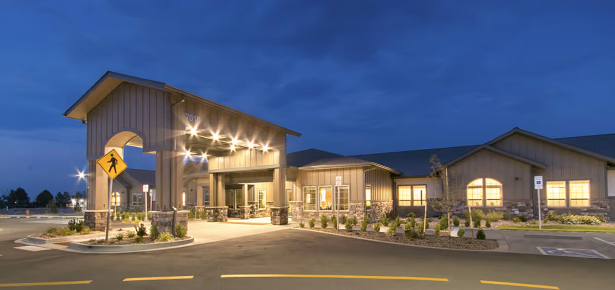 Exterior view of Windsong at Northridge facility at dusk, showing a well-lit entrance with a covered drop-off area, stone and wood siding, landscaped surroundings, and a pedestrian crossing sign in the foreground.