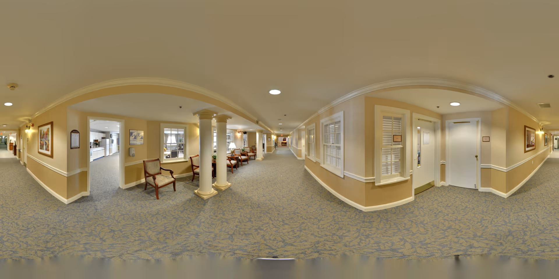 Wide panoramic view of a carpeted hallway in a senior living facility with beige walls and white trim. The hallway features several wooden chairs with cushions, decorative columns, framed artwork on the walls, and windows with white shutters. The corridor extends into the distance with multiple doors and ceiling lights.