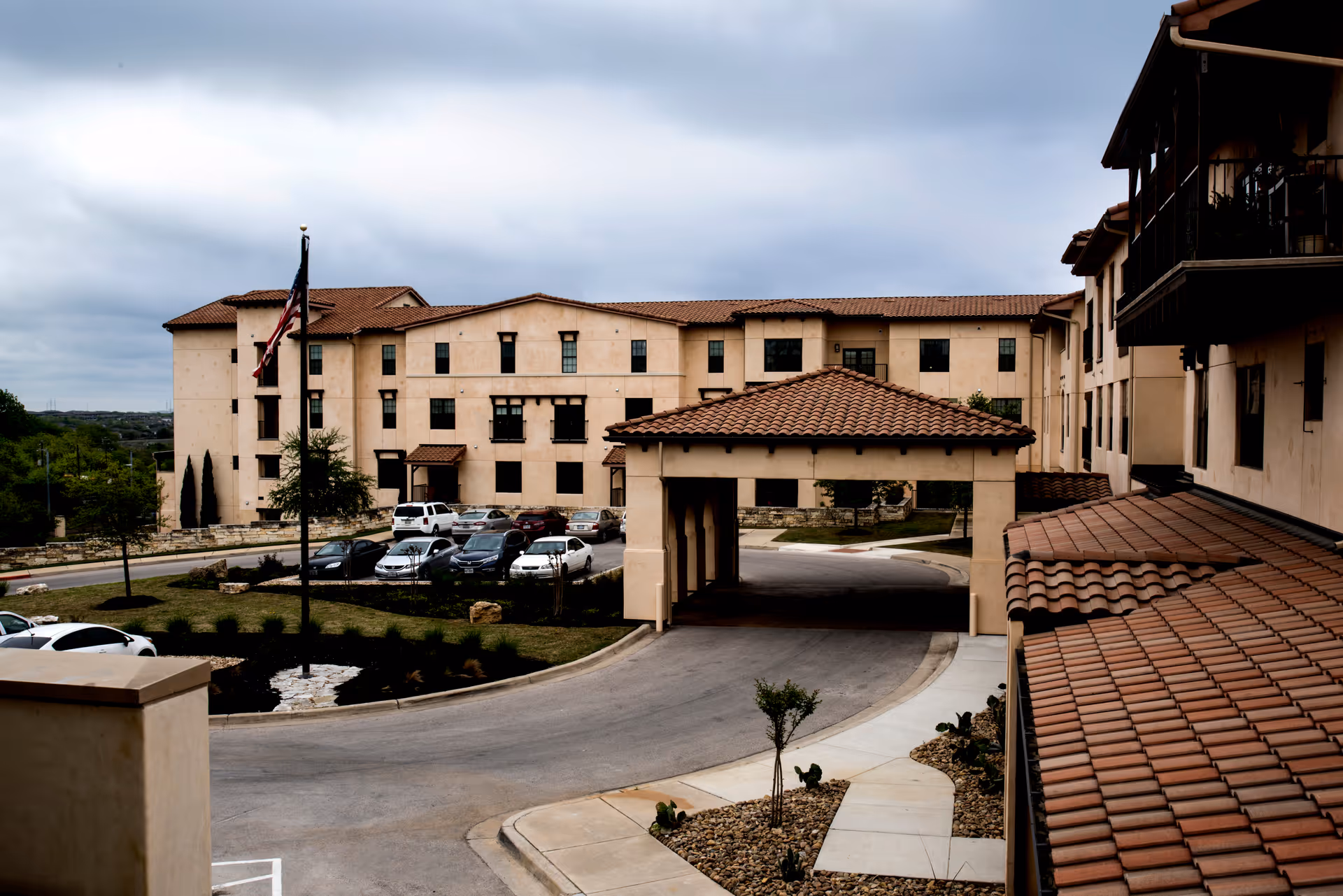 Exterior front view of a multi-story senior living building with a covered entrance, parking lot, and an American flag.