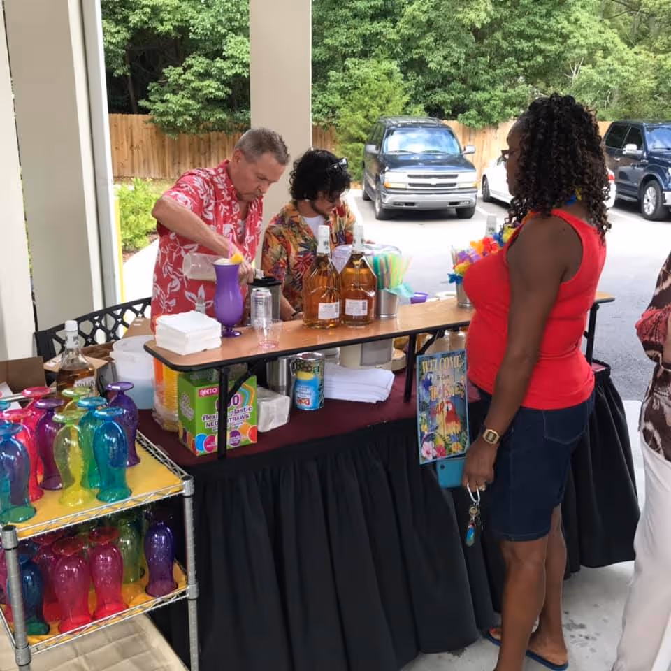 Three people at an outdoor bar setup with colorful plastic glasses and bottles of liquor. One man in a red floral shirt is pouring a drink, another person in a floral shirt is behind the bar, and a woman in a red sleeveless top and denim shorts is standing in front of the bar. There are cars and trees in the background.