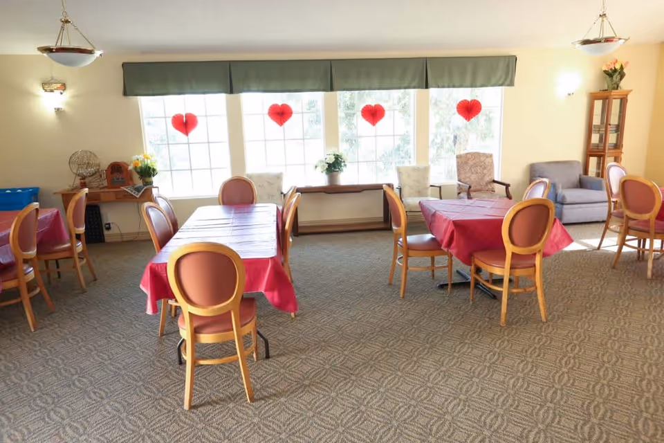 A bright dining room with several tables covered in red tablecloths and surrounded by wooden chairs with red cushions. Large windows with green valances let in natural light and have red heart decorations hanging. There are additional chairs and a small cabinet with flowers in the background.