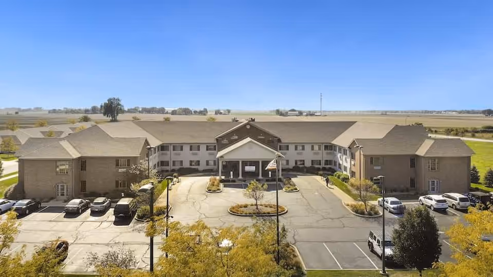 Front exterior view of Rittenhouse Village At Portage, a two-story senior living facility with a large driveway and parking area in front, surrounded by open fields under a clear blue sky.