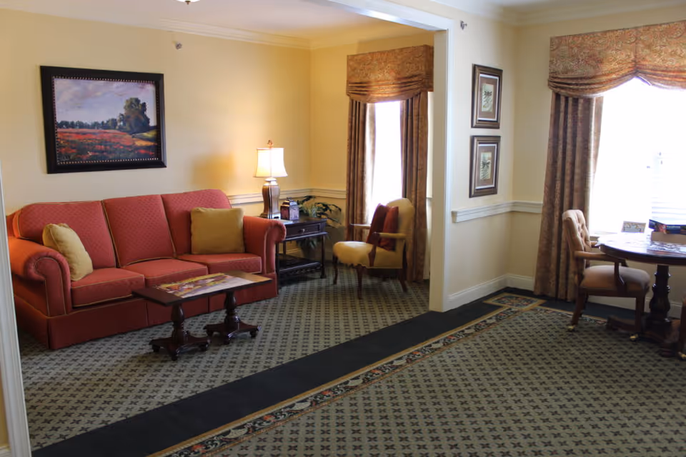 A cozy living room with a red sofa adorned with yellow cushions, a wooden coffee table, a side table with a lamp, and a yellow armchair near a window with patterned curtains. The room has a patterned carpet and framed artwork on the walls.