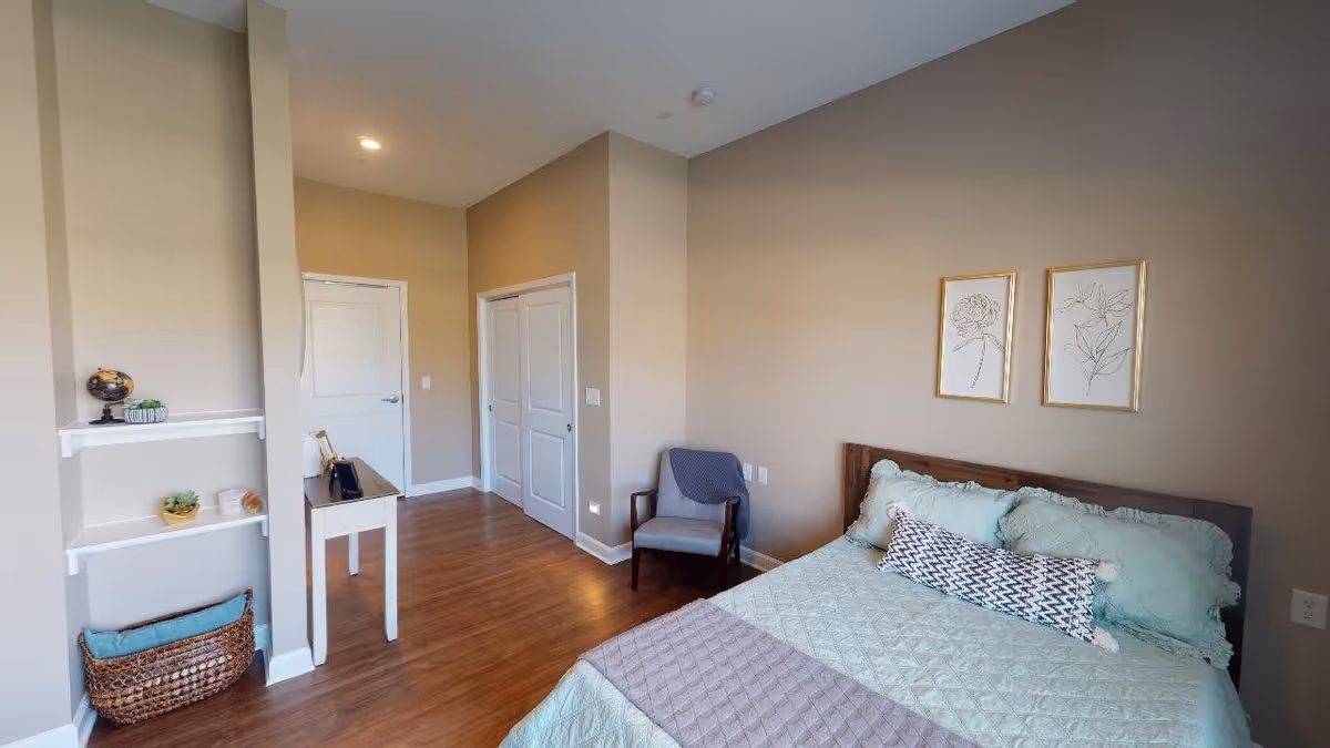 A cozy bedroom with a wooden bed featuring light green bedding and decorative pillows. Two framed botanical line art prints hang above the bed. There is a gray armchair with a blanket draped over it in the corner. The room has light beige walls, wooden flooring, a white door, a closet with double doors, and a small white table with decorative items. Shelves with small plants and a basket are visible on the left side.