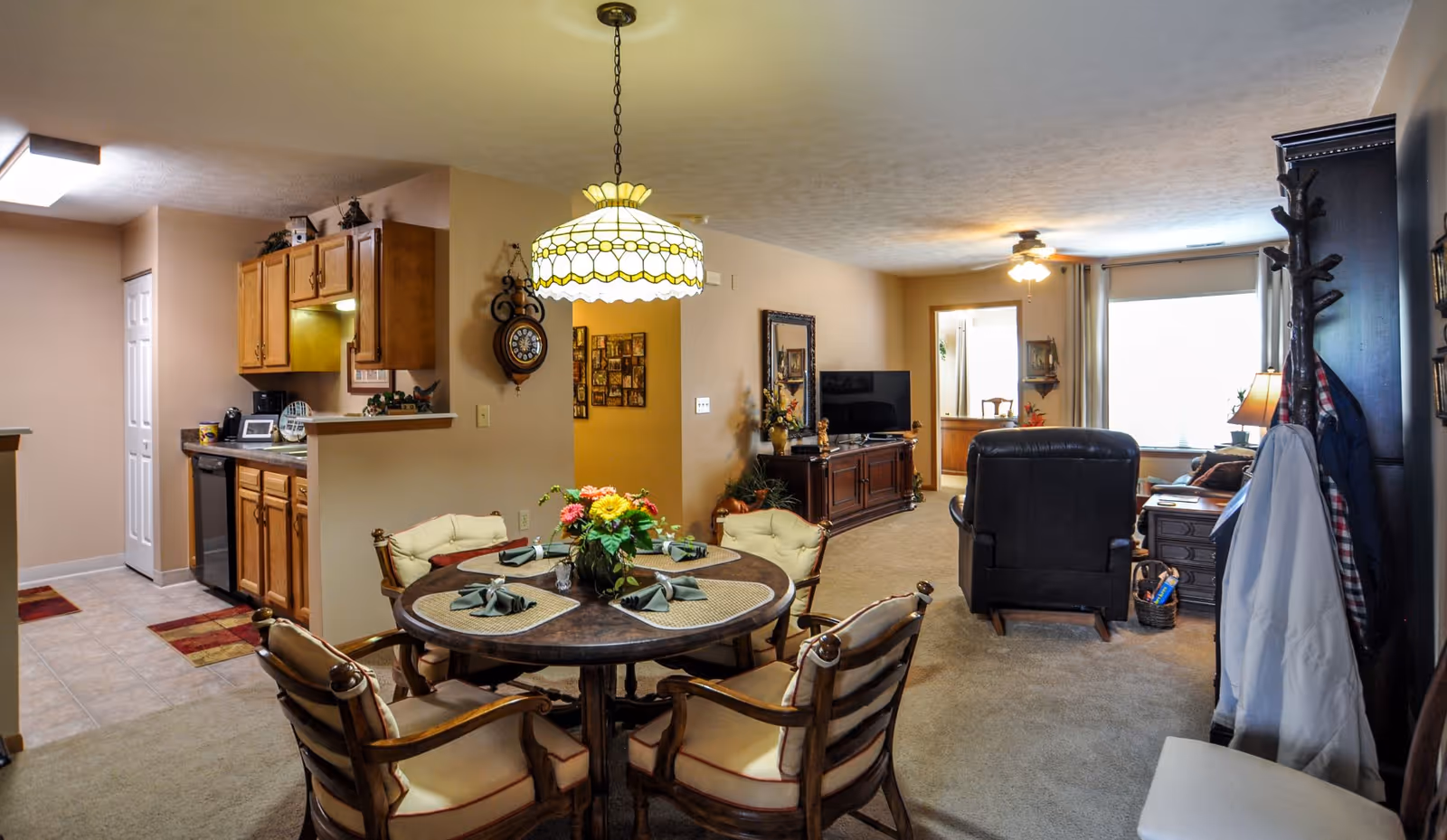 A cozy living and dining area in a senior living facility. The dining area features a round wooden table with four cushioned chairs, set with placemats, napkins, and a floral centerpiece. Adjacent to the dining area is a small kitchen with wooden cabinets and tiled flooring. The living room has a large window with curtains, a TV on a wooden stand, a recliner chair, and a coat rack with hanging clothes. The space is warmly lit with a ceiling light and a hanging lamp above the dining table.