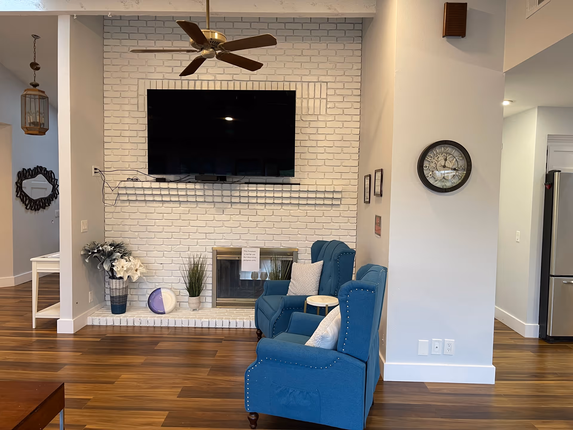 A cozy living room area with two blue armchairs facing a white brick fireplace. Above the fireplace is a large flat-screen TV mounted on the wall. There are decorative plants and a clock on the wall to the right. The room has wooden flooring and a ceiling fan with wooden blades.