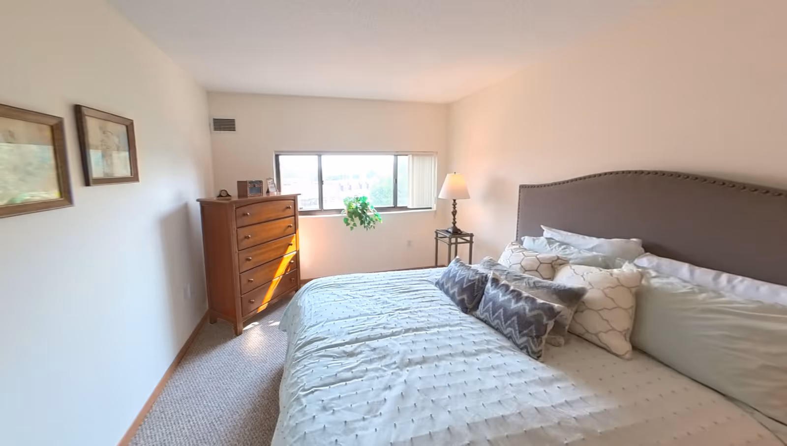 A bright bedroom with a large bed featuring multiple pillows and a gray upholstered headboard. There is a wooden chest of drawers near a window with vertical blinds, a small side table with a lamp, and two framed pictures on the wall. A green plant hangs in front of the window.