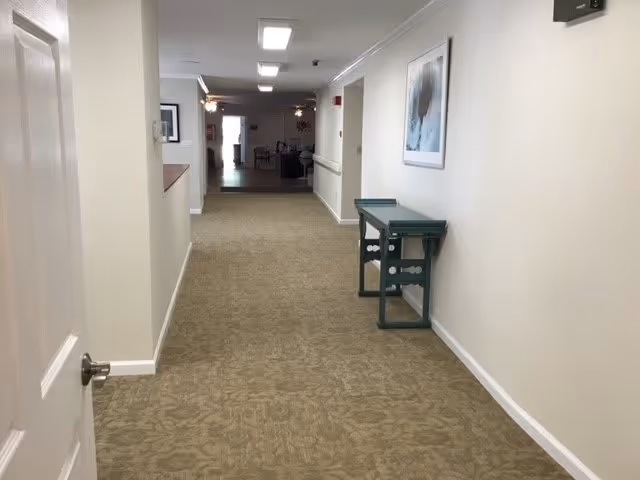 A long indoor hallway with beige patterned carpet and white walls. On the right side, there is a green console table and a framed picture hanging above it. The hallway leads to a more open area with furniture visible in the distance.