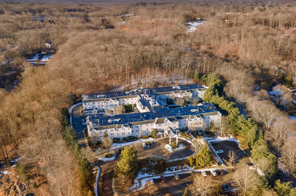 Aerial view of Monarch Spring Meadows, a large multi-wing senior living facility surrounded by leafless trees in a wooded area with patches of snow on the ground and parking lots with several cars.