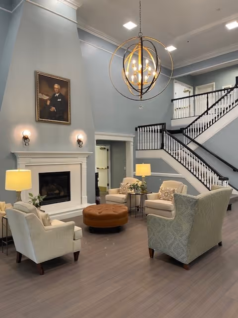 A spacious living room area with light blue walls and high ceilings featuring a modern chandelier. The room includes a white fireplace with two wall sconces and a portrait above it. There are four upholstered armchairs arranged around a round brown ottoman, with two table lamps on side tables. A staircase with white spindles and dark handrails leads to an upper floor.