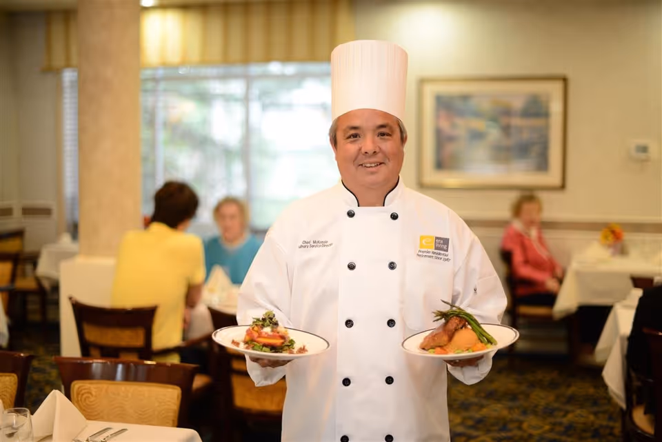 A chef wearing a white uniform and tall chef hat is smiling and holding two plates of food in a dining room with tables, chairs, and people seated in the background.