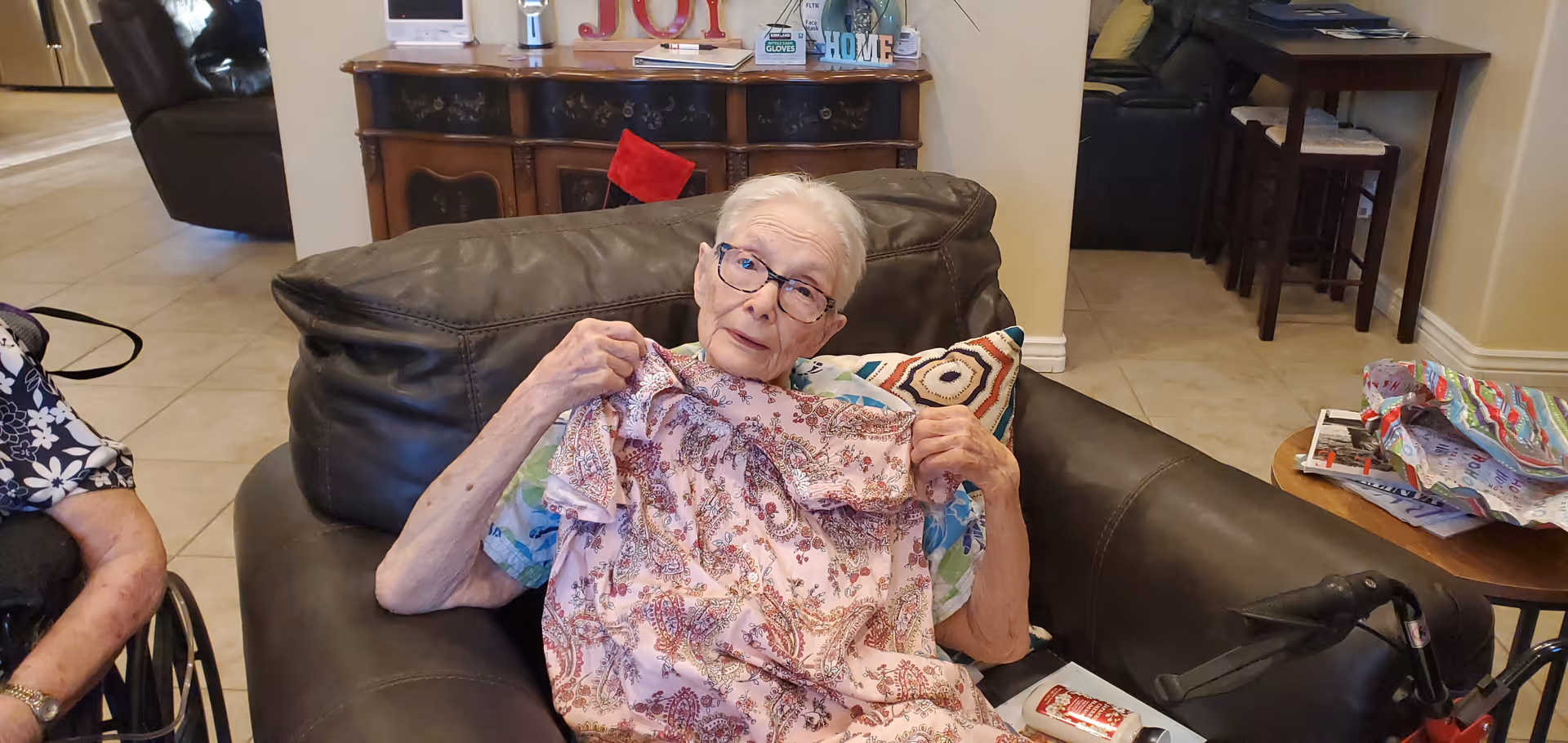 An elderly woman sitting on a dark brown leather couch in a living room, holding up a pink patterned garment. The room has tiled floors, a wooden sideboard with decorative items including a sign that says 'JOY', and a small table with some papers and a walker nearby.