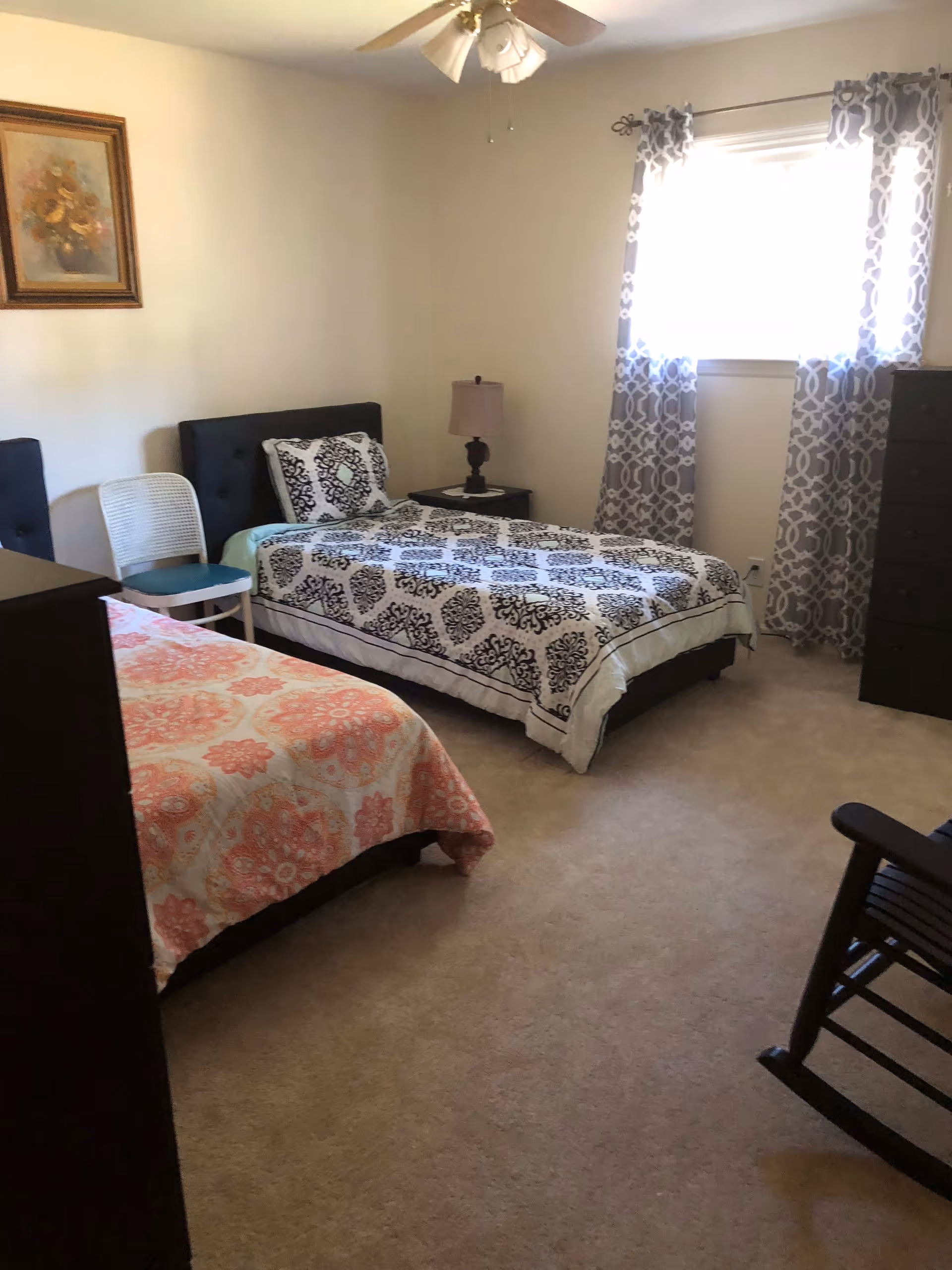 A bedroom with two twin beds, one with a black and white patterned bedspread and the other with a pink and white patterned bedspread. There is a white chair between the beds, a nightstand with a lamp, a dresser, a rocking chair, a window with patterned curtains, and a ceiling fan with lights. A framed floral painting hangs on the wall.