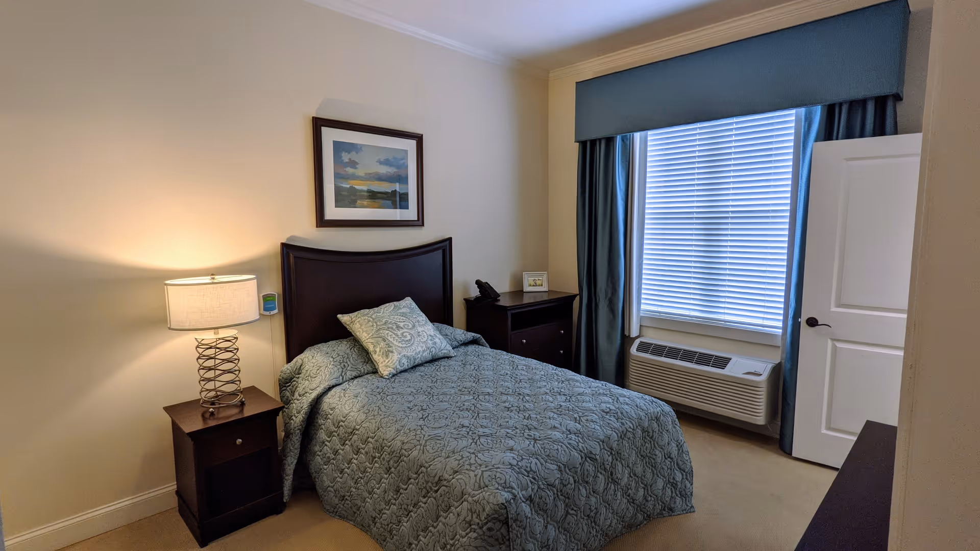 A neatly made single bed with a patterned blue-green bedspread and matching pillow in a small bedroom. The room features a wooden headboard, a nightstand with a lamp, a framed landscape picture on the wall, a window with blinds and blue curtains, an air conditioning unit below the window, and a partially open white door.