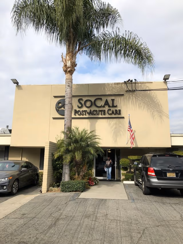 A building entrance of SoCal Post Acute Care with a palm tree and parked cars.
