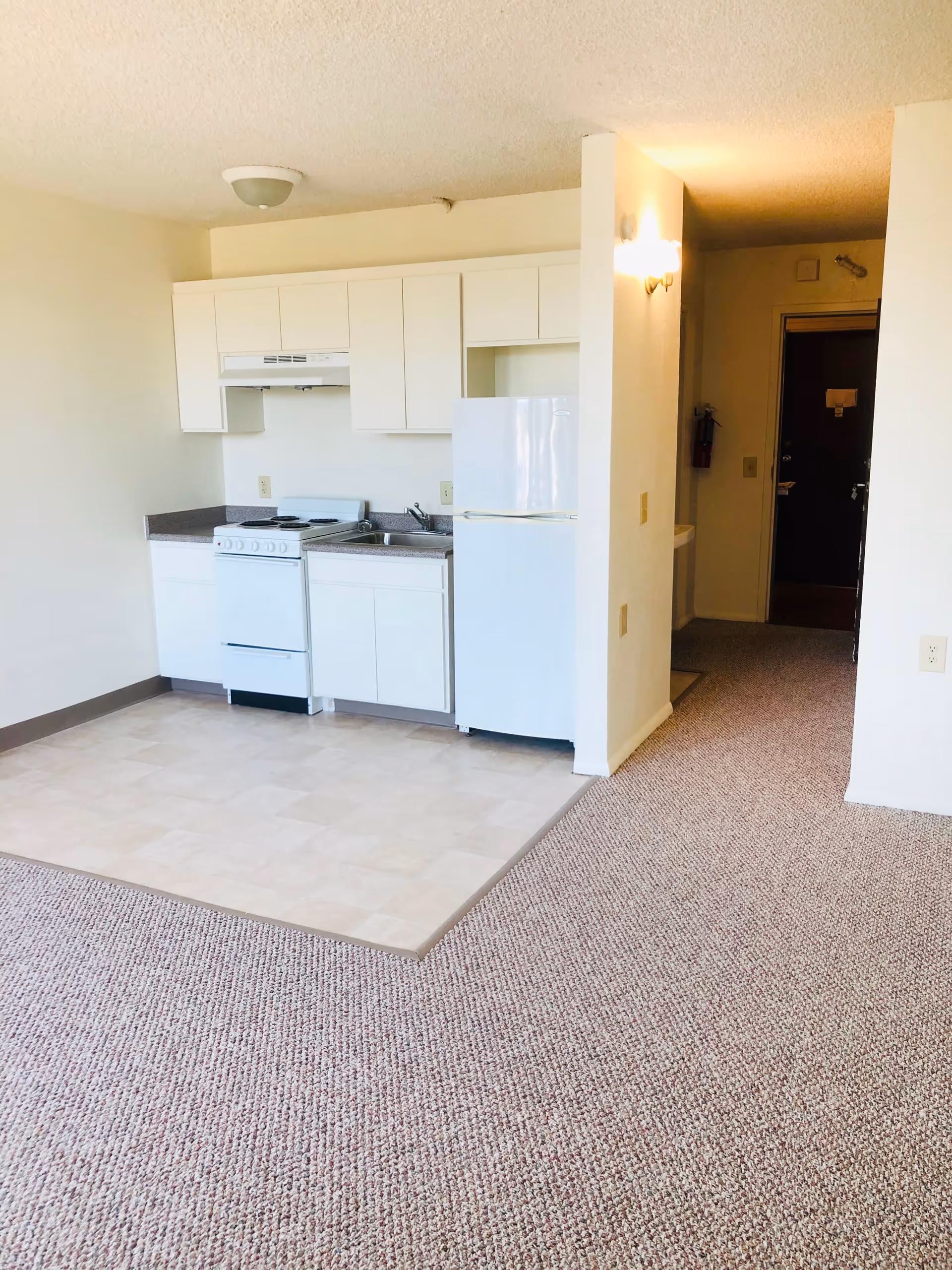 Small kitchen area in a senior living facility with white cabinets, a white stove, a white refrigerator, and a sink. The kitchen floor is tiled, and the surrounding area is carpeted. There is a hallway leading to a dark door with a fire extinguisher mounted on the wall.