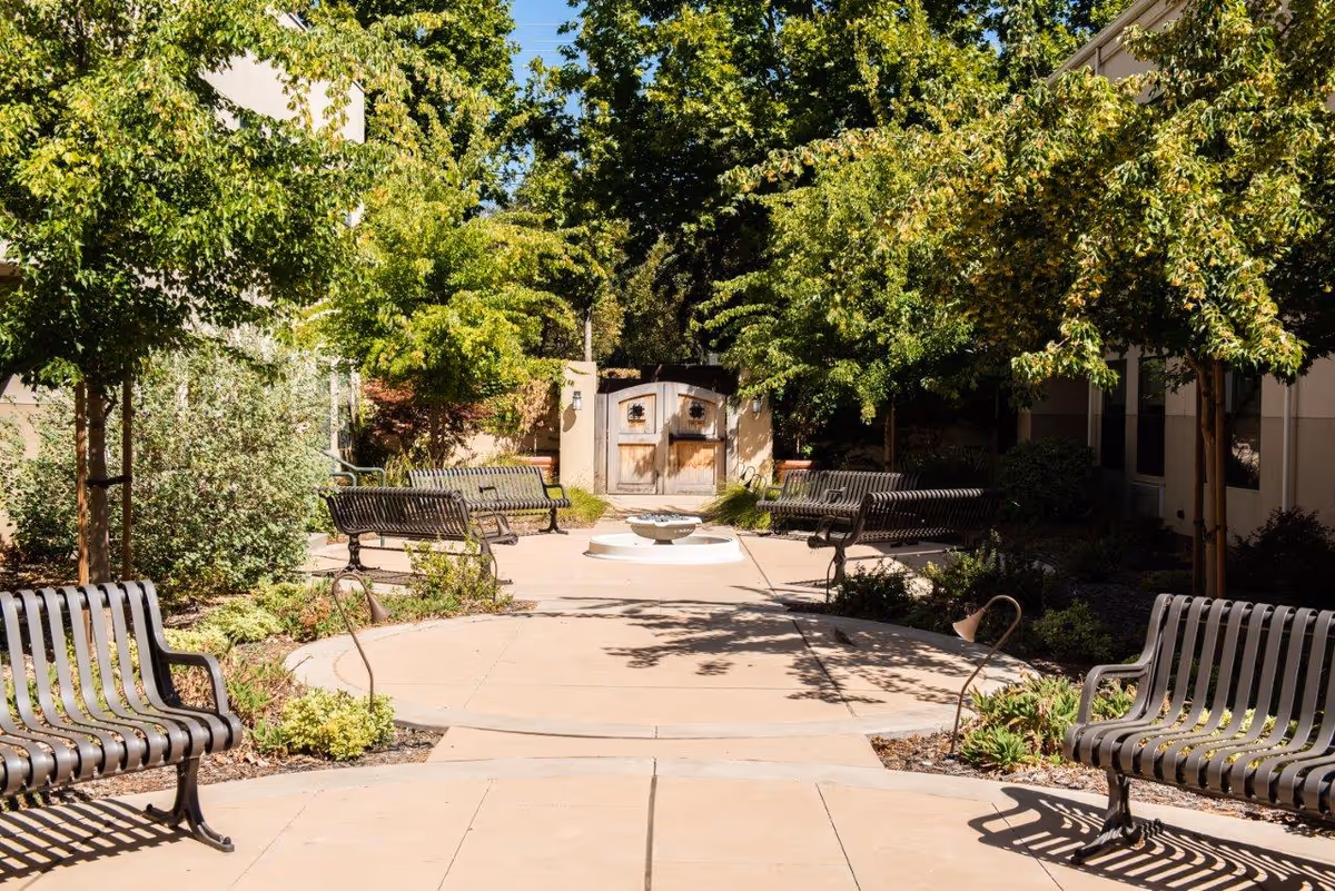 Sunlit courtyard with metal benches arranged around a small central fountain, surrounded by trees and landscaping in front of a wooden gate.